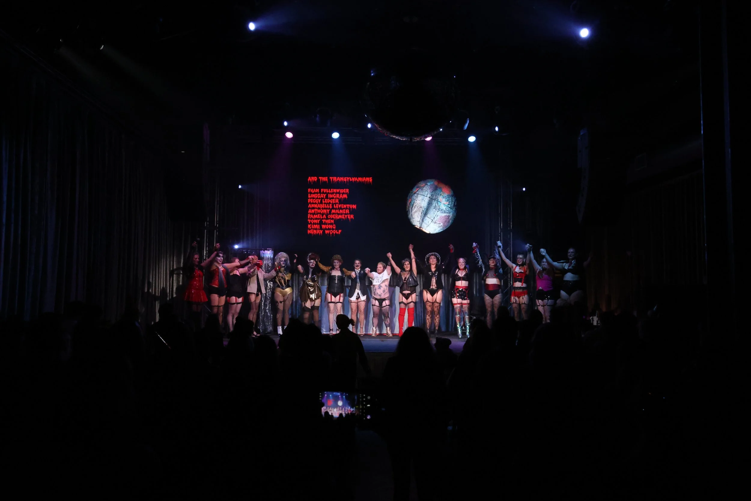Group of performers on stage in colorful costumes, holding hands and bowing, with a red digital display above them showing cast and crew names.