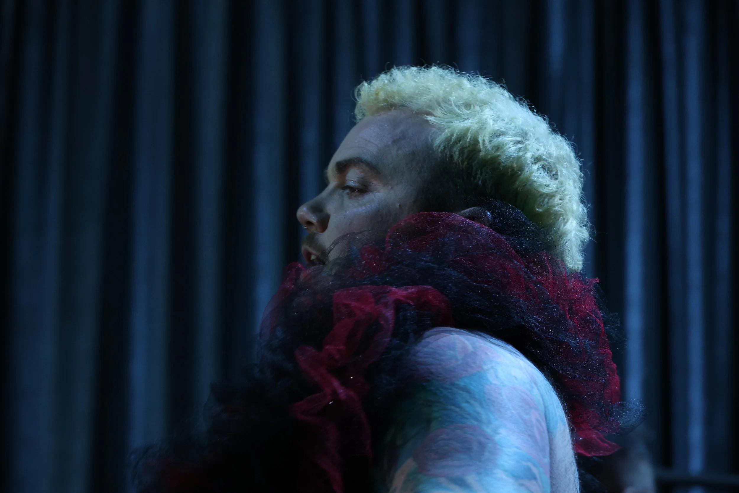 A person with blonde, curly hair in profile view, wearing a colorful, textured garment, against a dark, striped background.