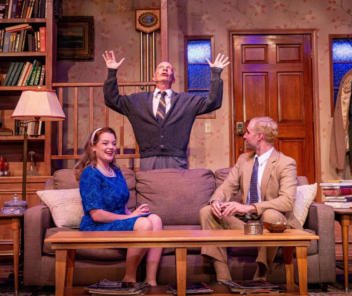A theatrical scene with three actors on stage, featuring a man standing with his hands raised, a smiling woman in a blue dress sitting on a couch, and a man in a tan suit sitting beside her. The setting is a living room with bookshelves, a lamp, and 