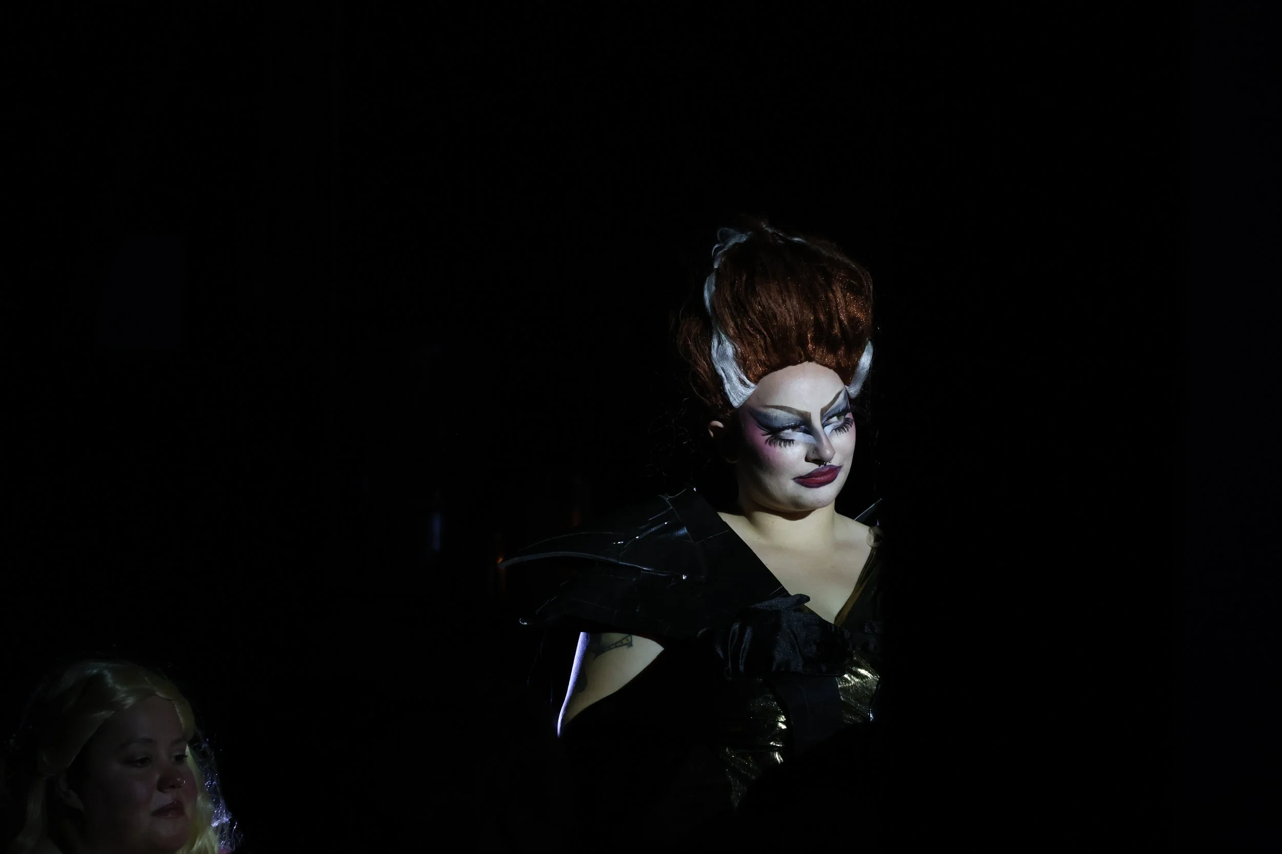 A person with elaborate makeup and red hair styled in an updo, wearing a dark costume, stands against a black background, illuminated by a spotlight.