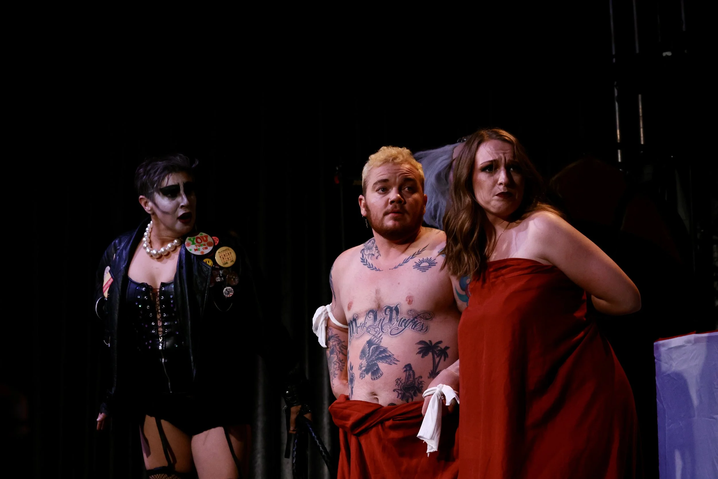 Three people are on stage in a theatrical performance. The person on the left is a woman with dark makeup, wearing a black leather jacket and accessories. The person in the middle is a shirtless man with tattoos and a towel wrapped around his waist, 