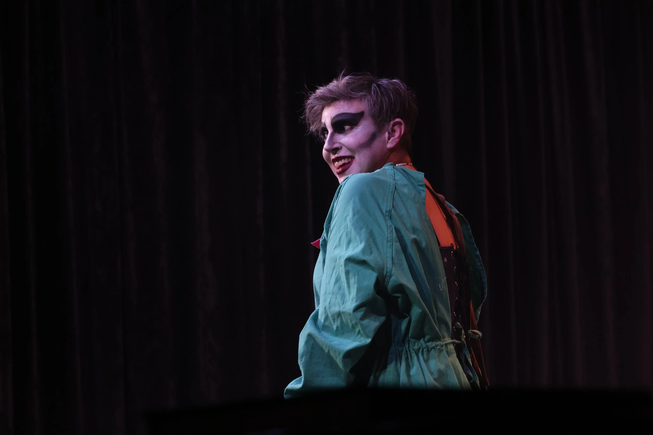 A person with theatrical makeup, dark eye makeup, and a green jacket, smiling and looking to the side, standing against a dark curtain background.
