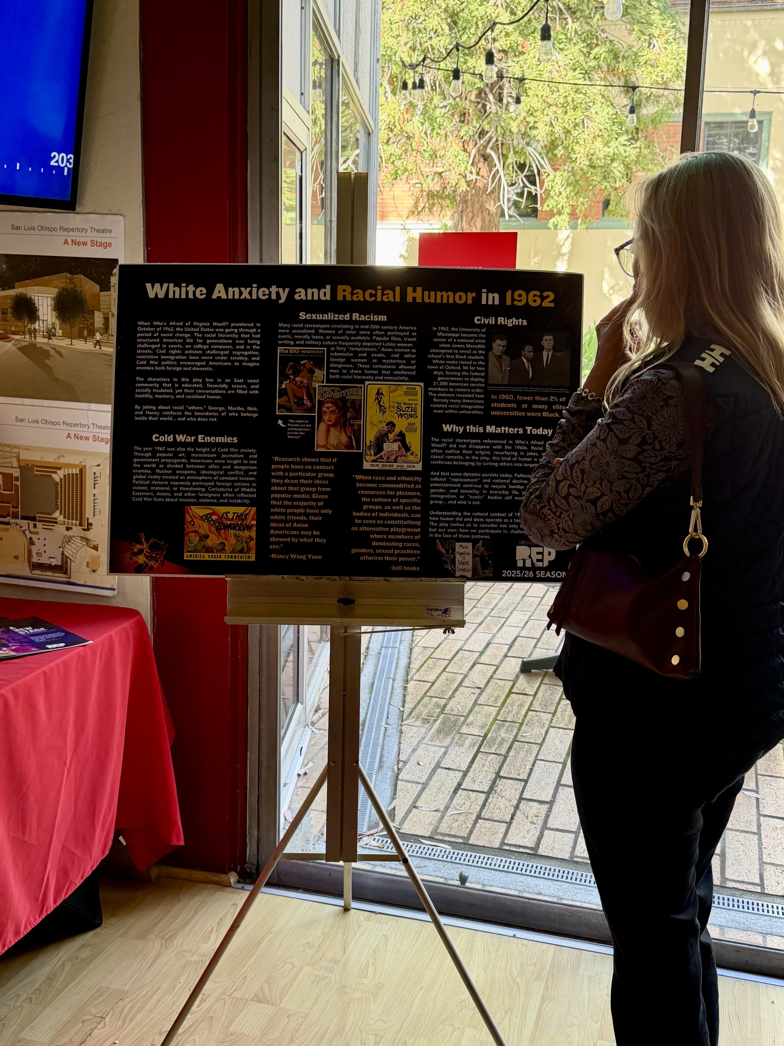 A woman with long blond hair, glasses, and a black outfit stands indoors near a large window, looking at an informational poster about racial topics from 1962, with trees and a building visible outside.