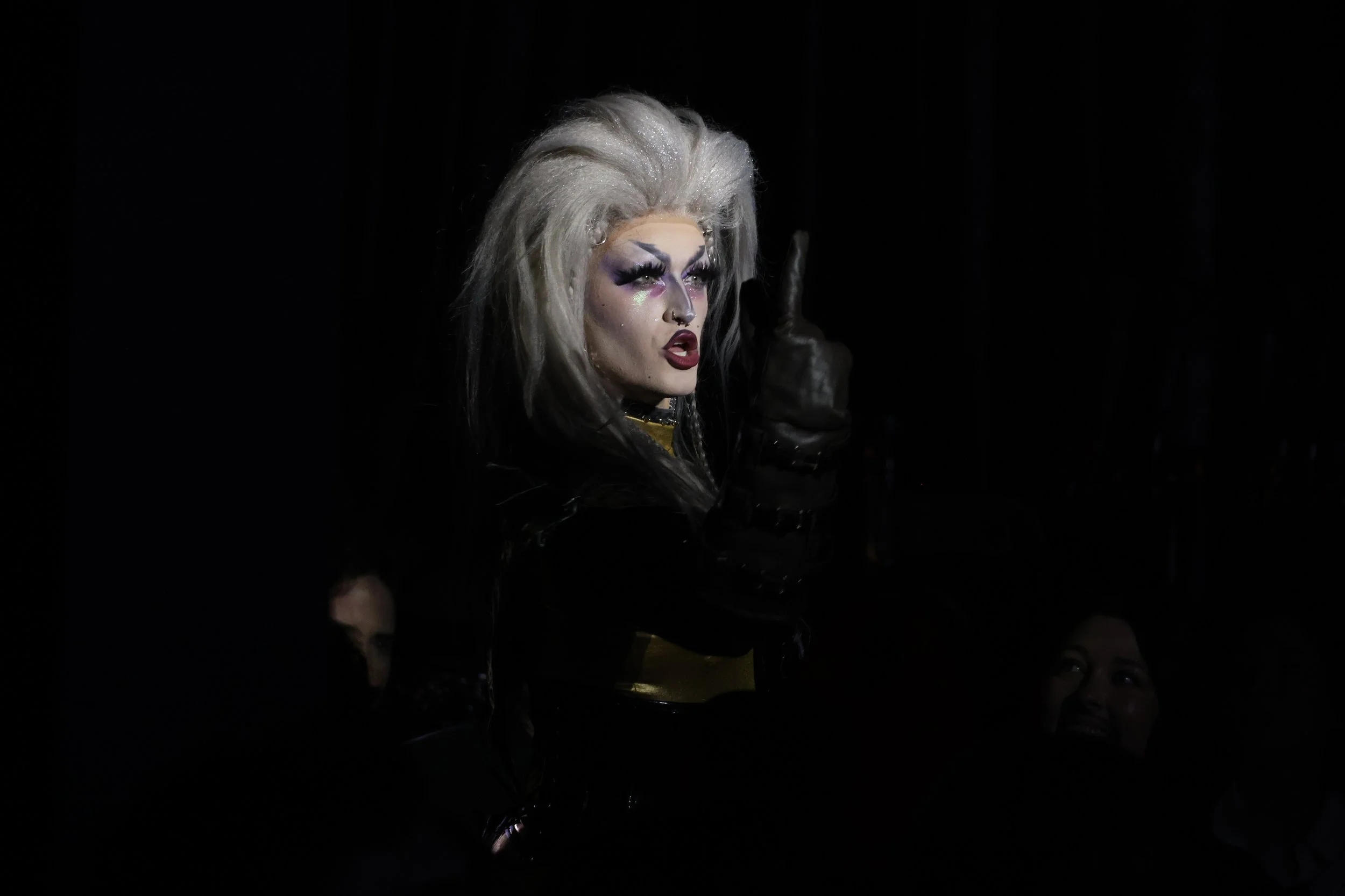 Person with platinum blonde hair styled in a voluminous manner, wearing dramatic eye makeup and dark lipstick, making a gesture with their hand. The background is dark, and part of another person's face is visible at the bottom right corner.