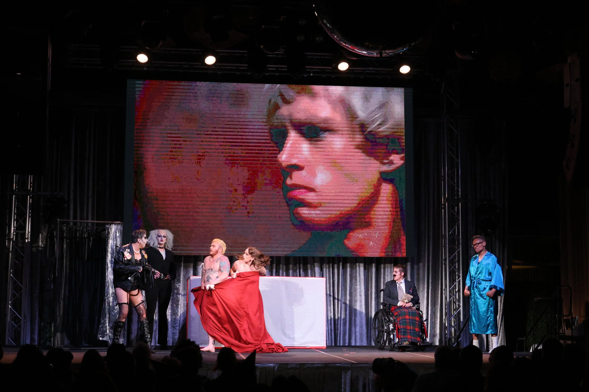 Stage scene with a large screen displaying a colorful portrait of a woman with short blonde hair. Several people are on stage, some in costumes, including a person in a wheelchair and a person in a shiny blue outfit. The stage has black curtains and 