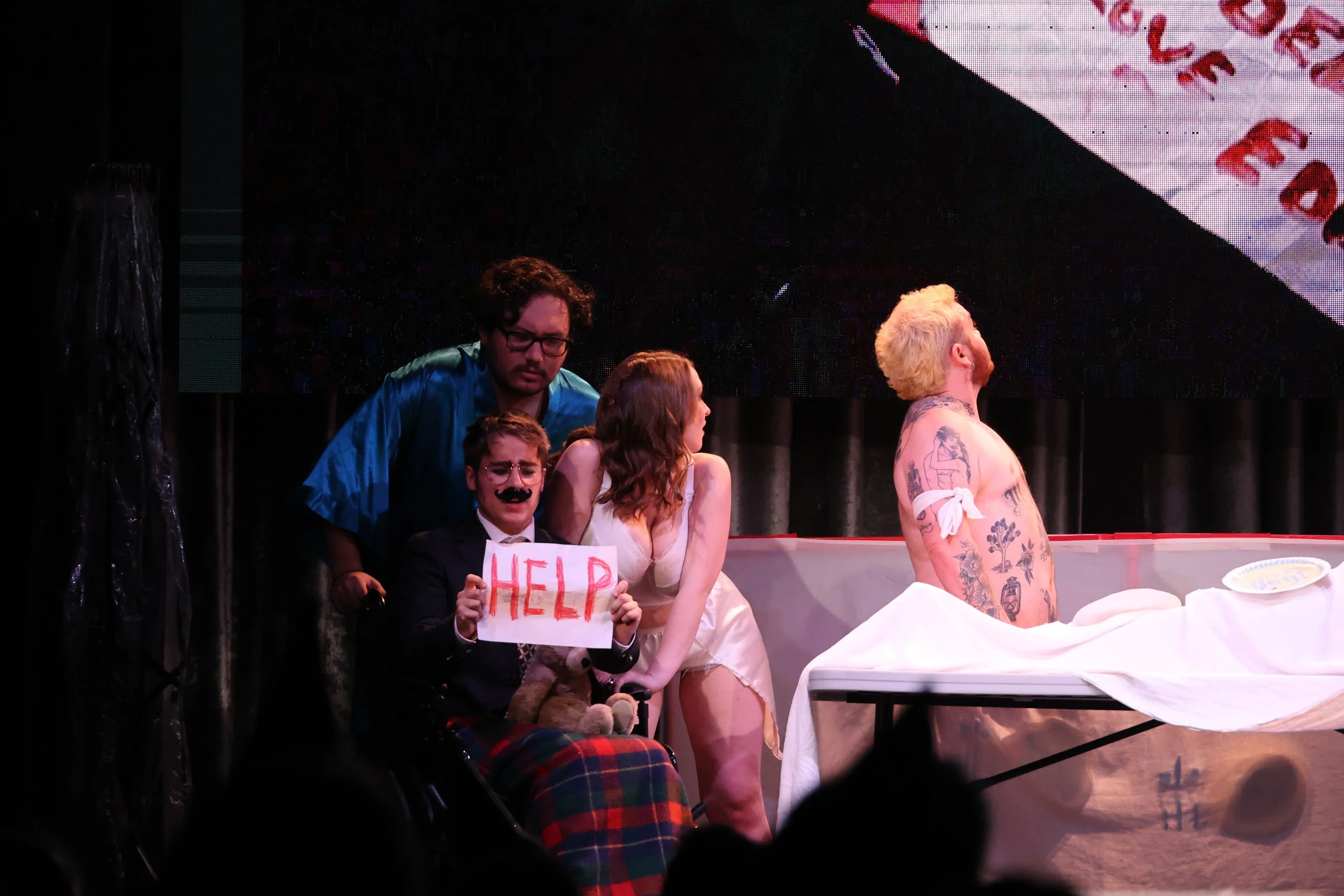 A theatrical scene with five actors, one holding a 'HELP' sign, another with a mustache and glasses, and a shirtless man with tattoos, sitting at a table covered with a white cloth.