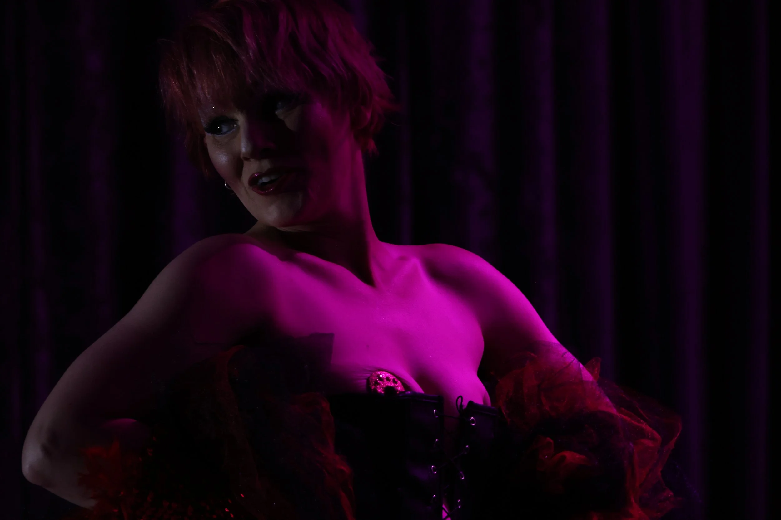 A woman with short red hair and glamorous makeup sitting topless on a bed of red roses, illuminated by purple and pink lighting with a dark background.