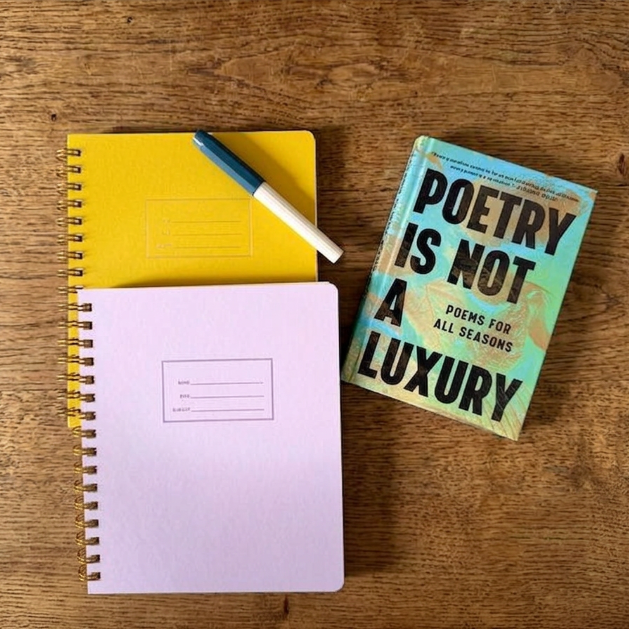 A yellow spiral notebook, a white and blue pen, a pink notebook, a paperback book titled "Poetry is not for everybody", all placed on a wooden surface.