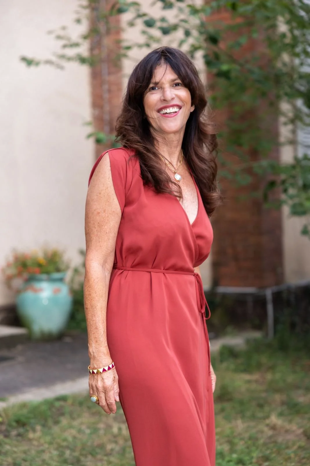 A woman with long brown hair smiling outdoors, wearing a red dress, jewelry, and a bracelet, with greenery and a brick building in the background.
