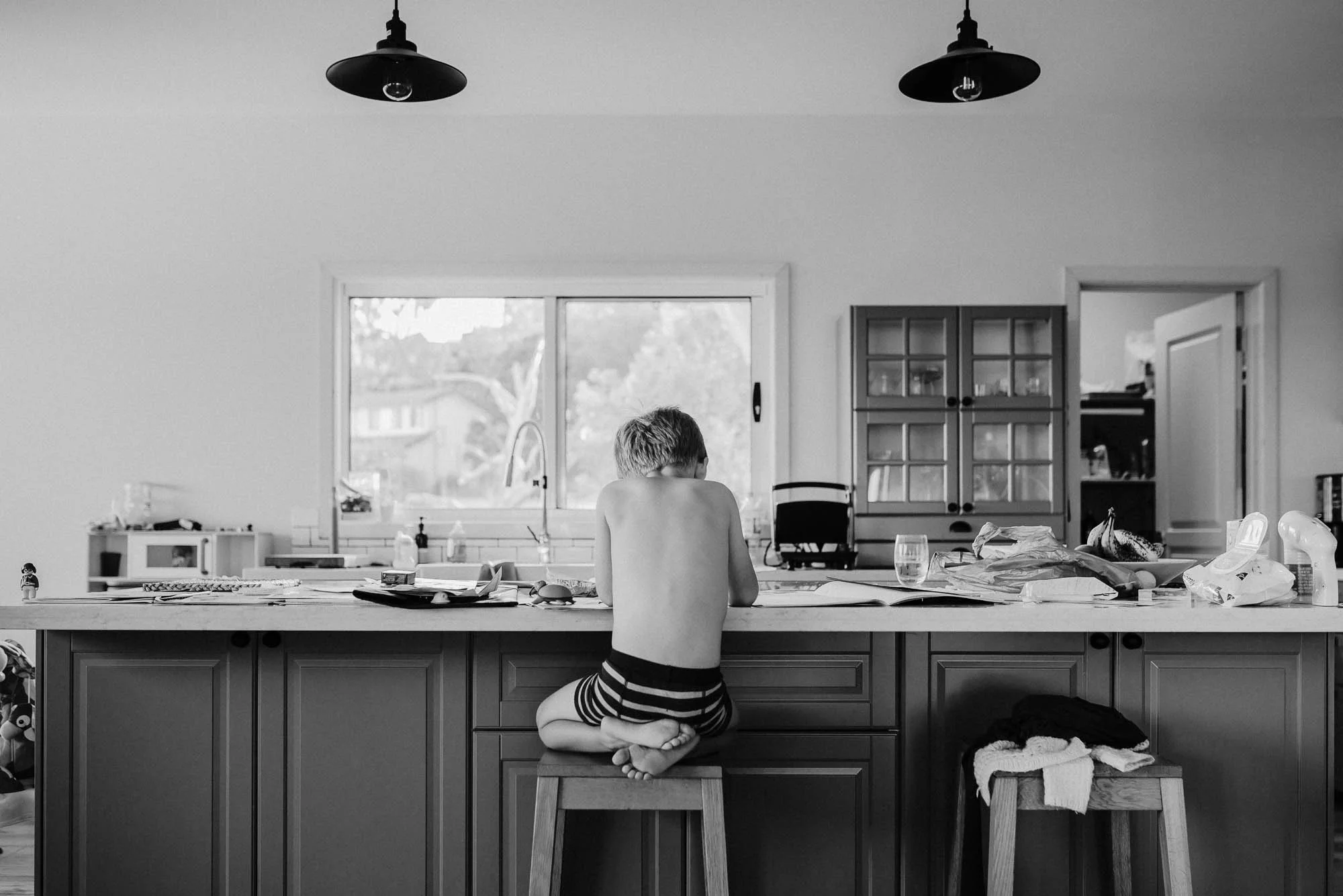 black_and_white_image_of_the_back_of_a_boy_sitting_on_his_feet_at_the_kitchen_counter.jpg