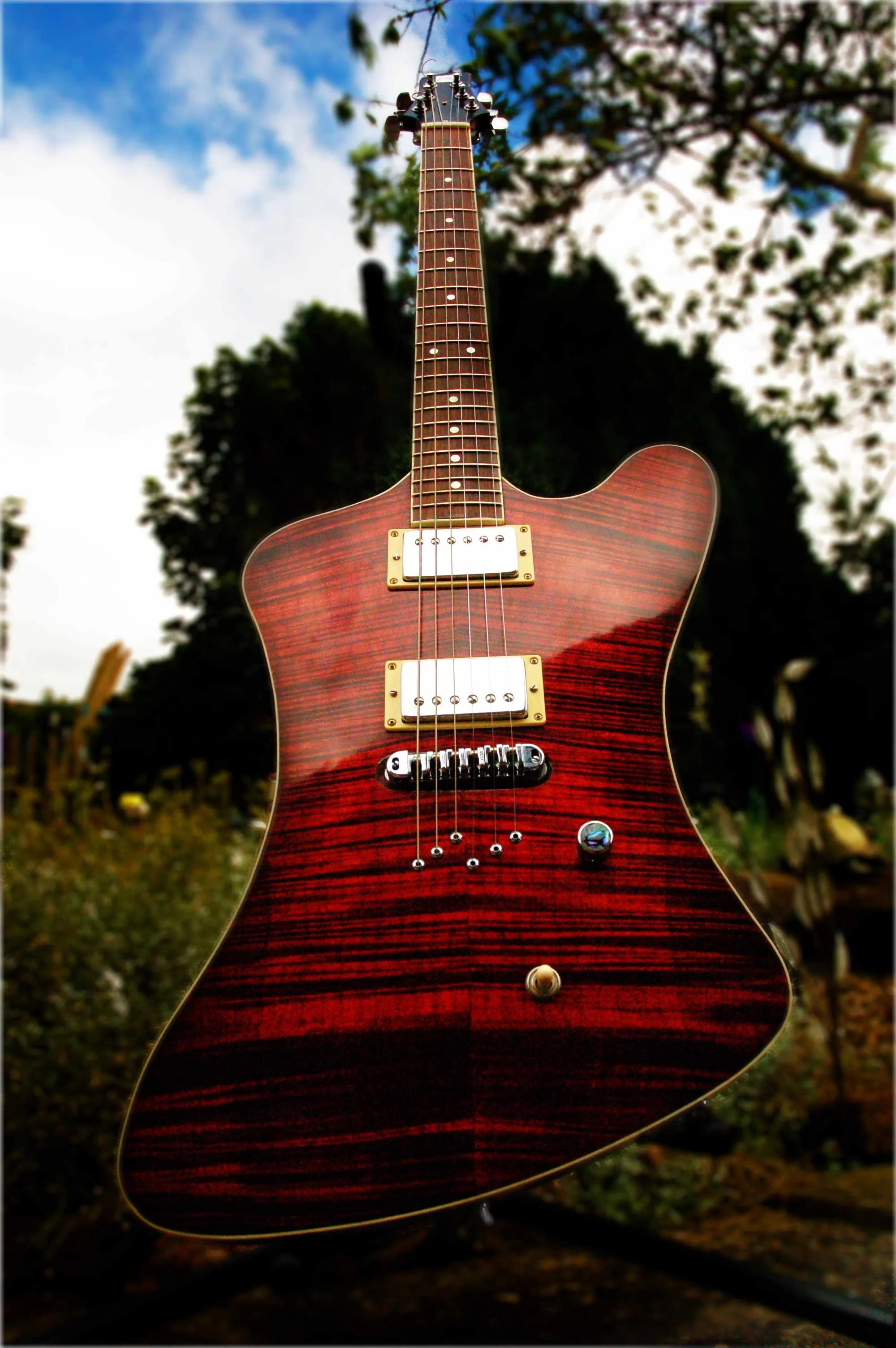 A close-up view of a red, wood-finish electric guitar with gold hardware, set outdoors against a background of trees and a partly cloudy sky.