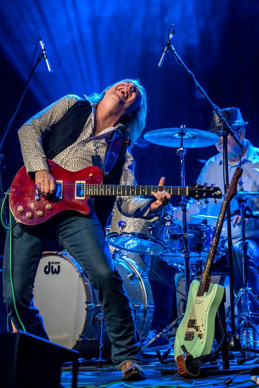 Musician playing a red electric guitar and singing on stage, with a drummer and another guitar visible in the background, under blue stage lighting.