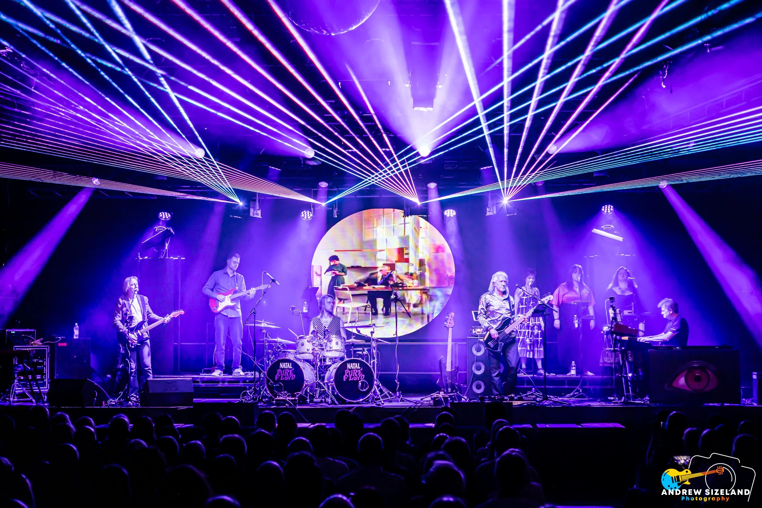 A live musical performance on stage with multiple musicians, including guitarists, a bassist, a drummer, and a keyboard player, under vibrant purple and blue laser lights and illuminated stage effects.