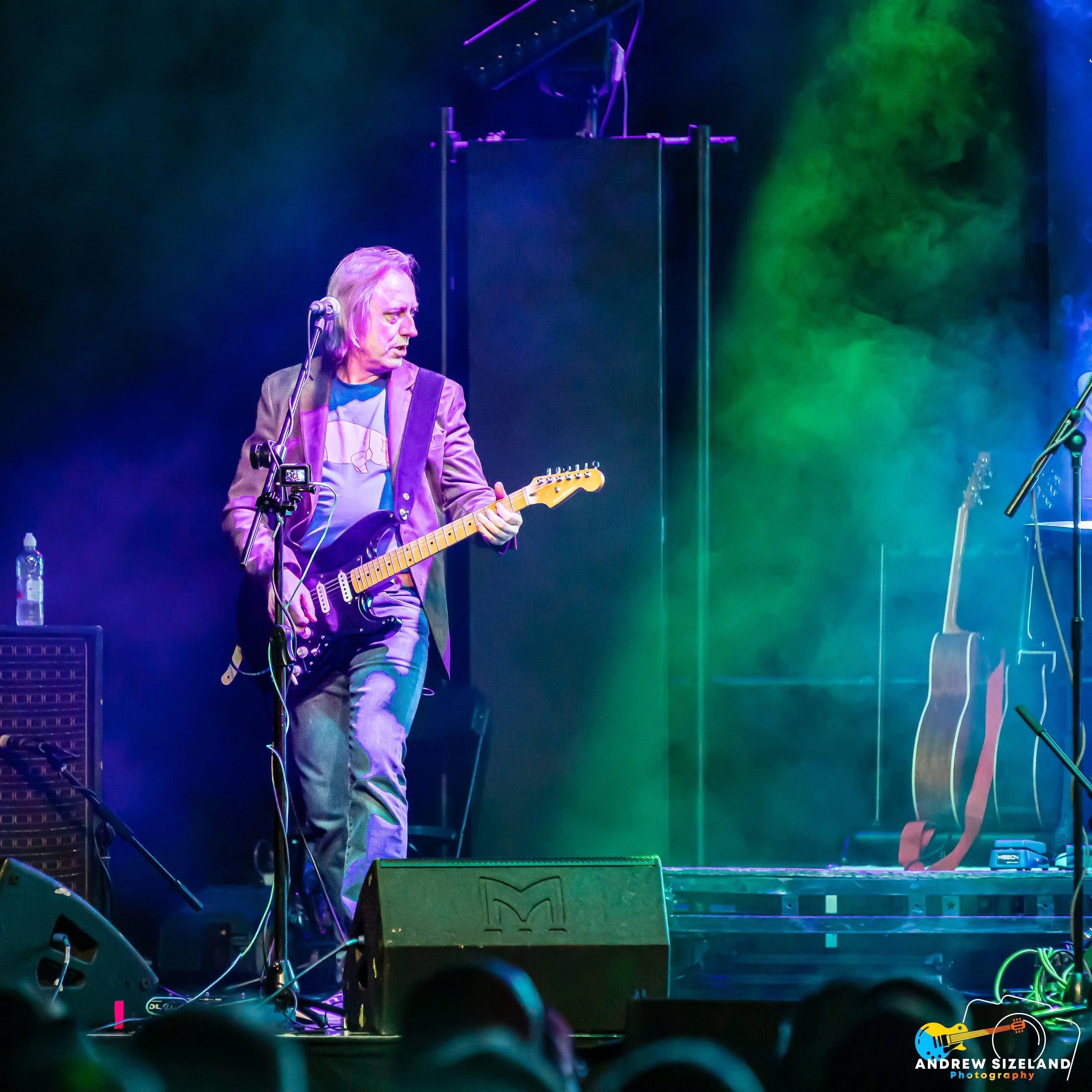 A man with long hair and a blazer playing an electric guitar on a stage with colorful stage lighting and fog effects, with music equipment and a microphone stand visible.