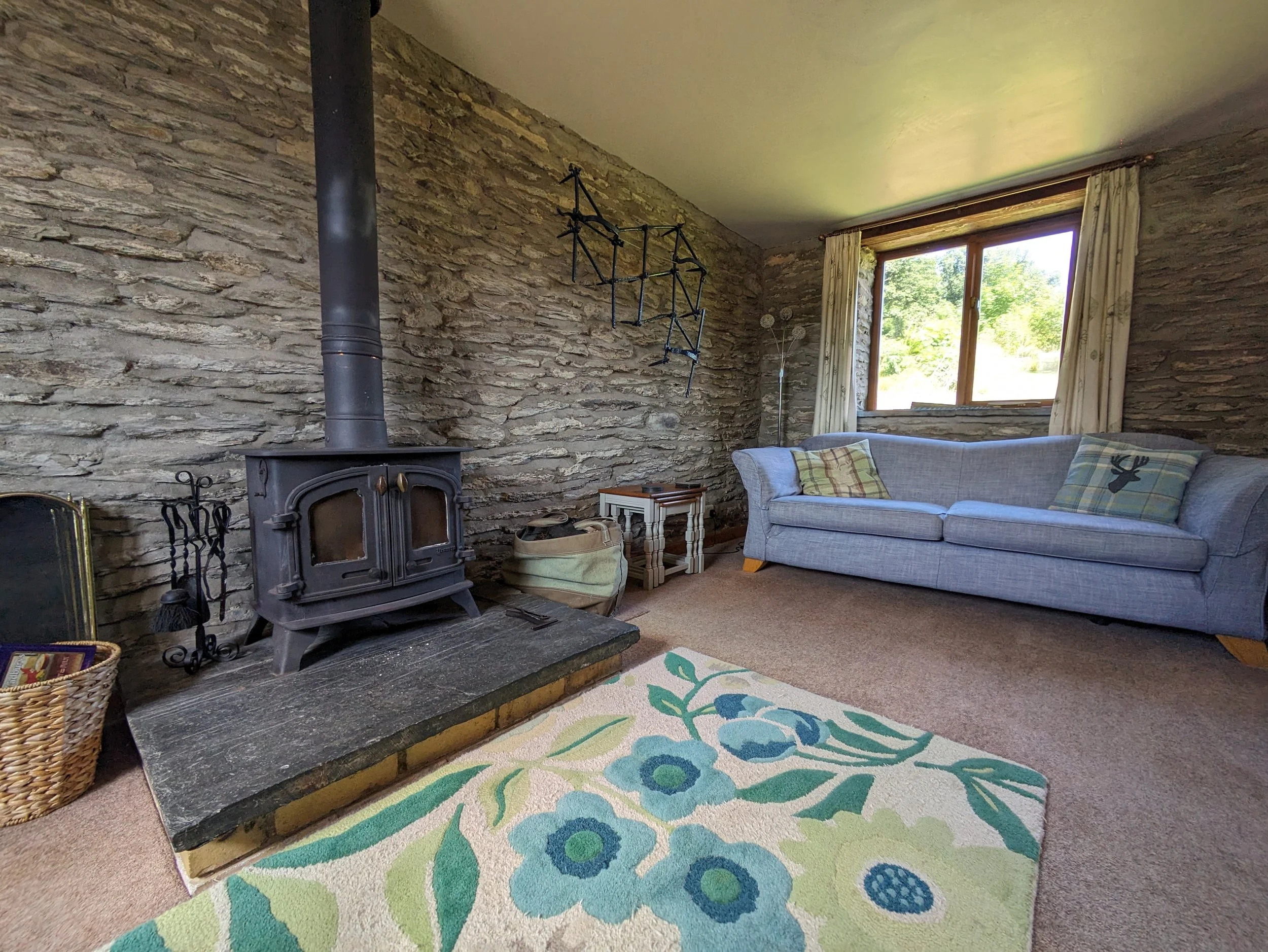 Living room with stone brick wall, wood stove, gray sofa with plaid pillows, window with cream curtains, colorful floral rug, and small side tables.