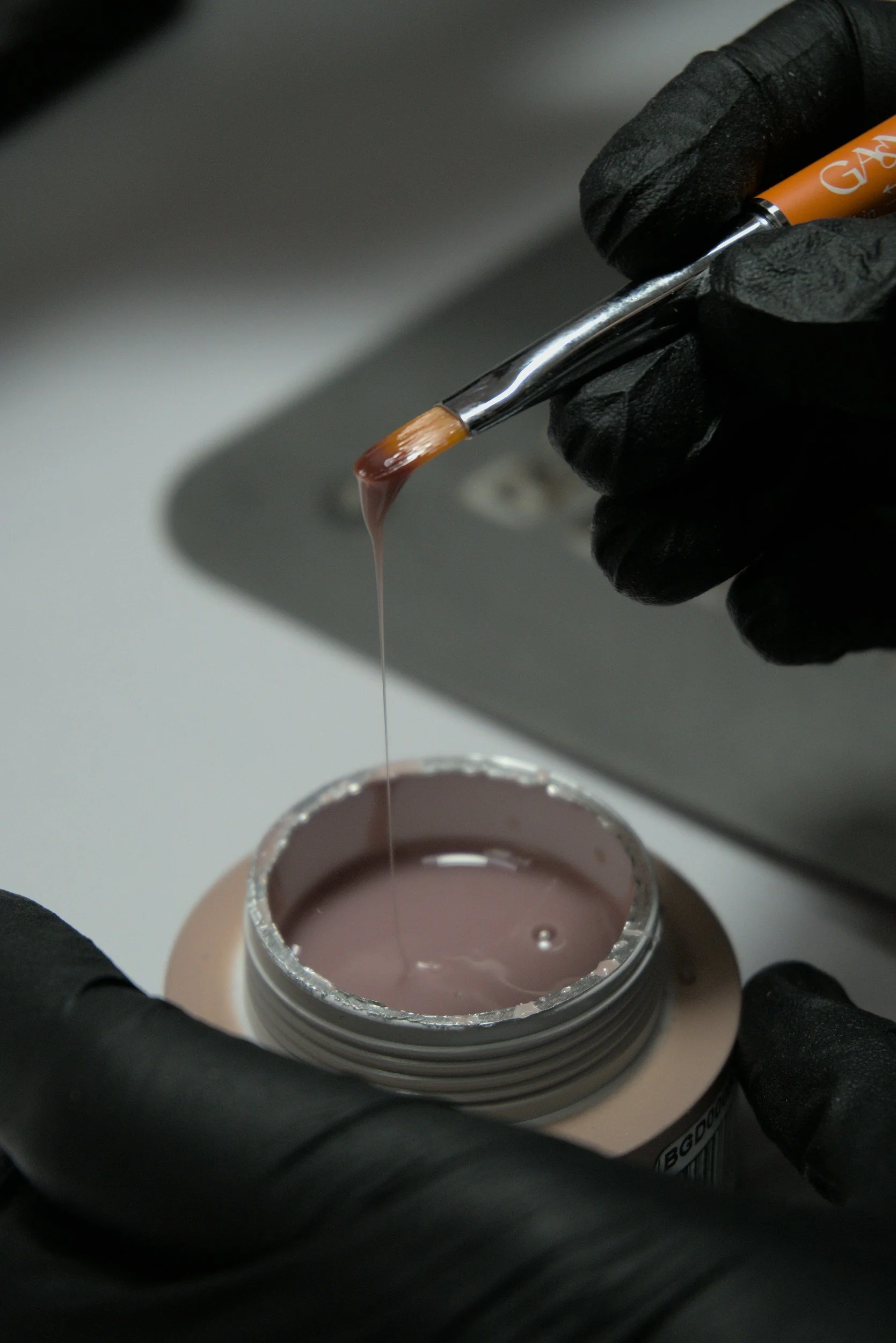 Close-up of a person wearing black latex gloves holding a small brush dipped in pink acrylic paint over a container filled with pink paint.