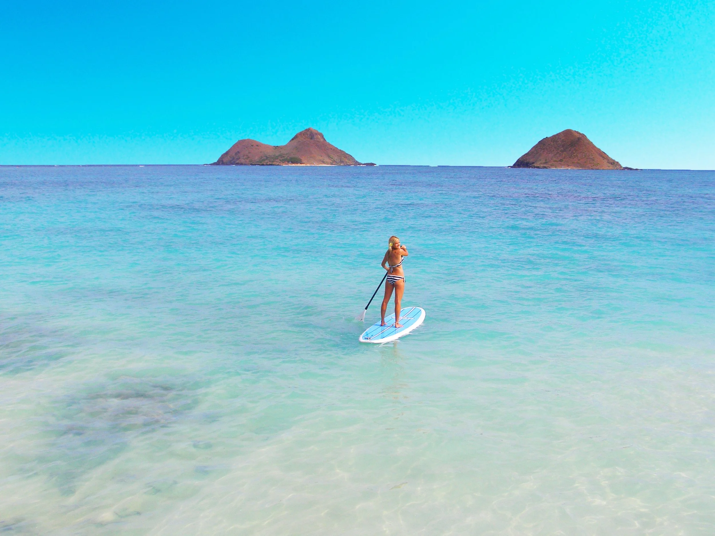 A woman standing on a paddleboard in clear blue waters with two small islands in the background.