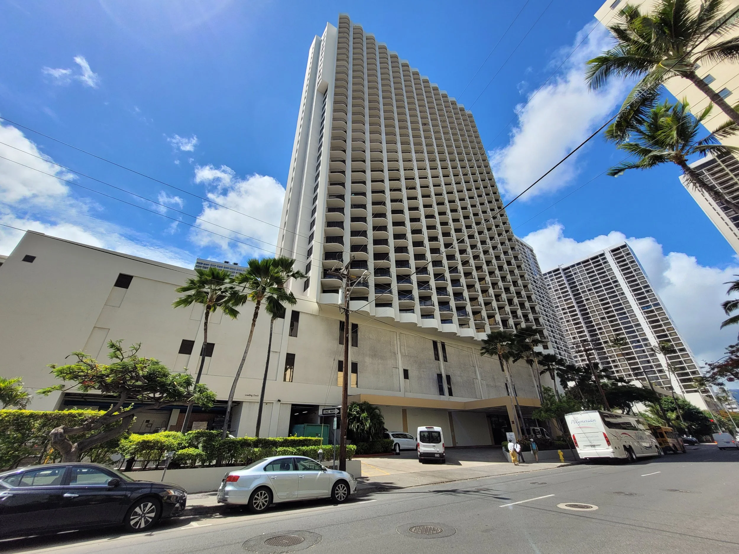 Tall white multi-story hotel building with curved balconies, palm trees, cars parked on the street, and a blue sky with clouds.