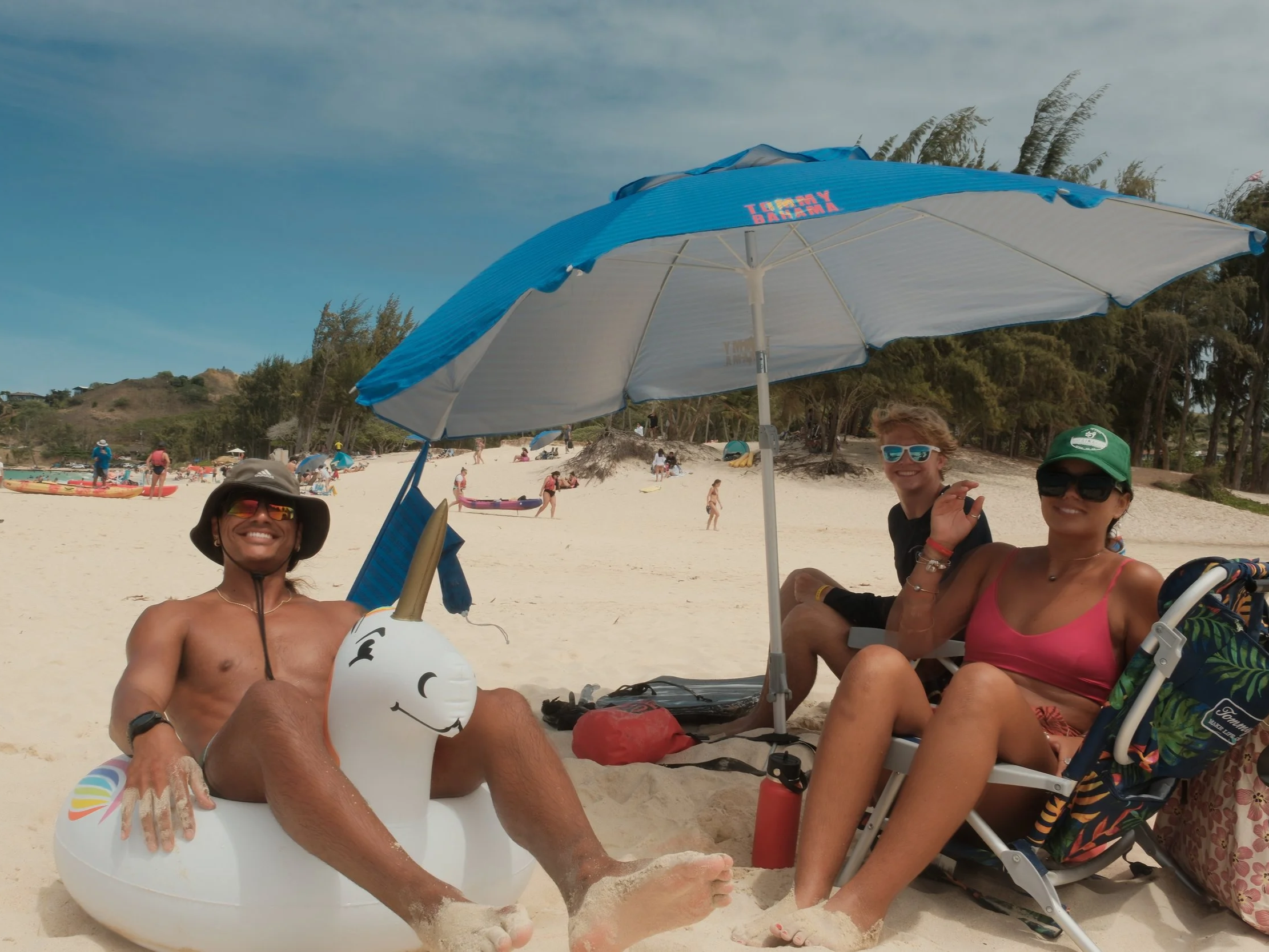 Three friends sitting under a blue umbrella at the beach, smiling and wearing sunglasses. One person is holding an inflatable unicorn, and others are relaxing on beach chairs with trees and other beachgoers in the background.