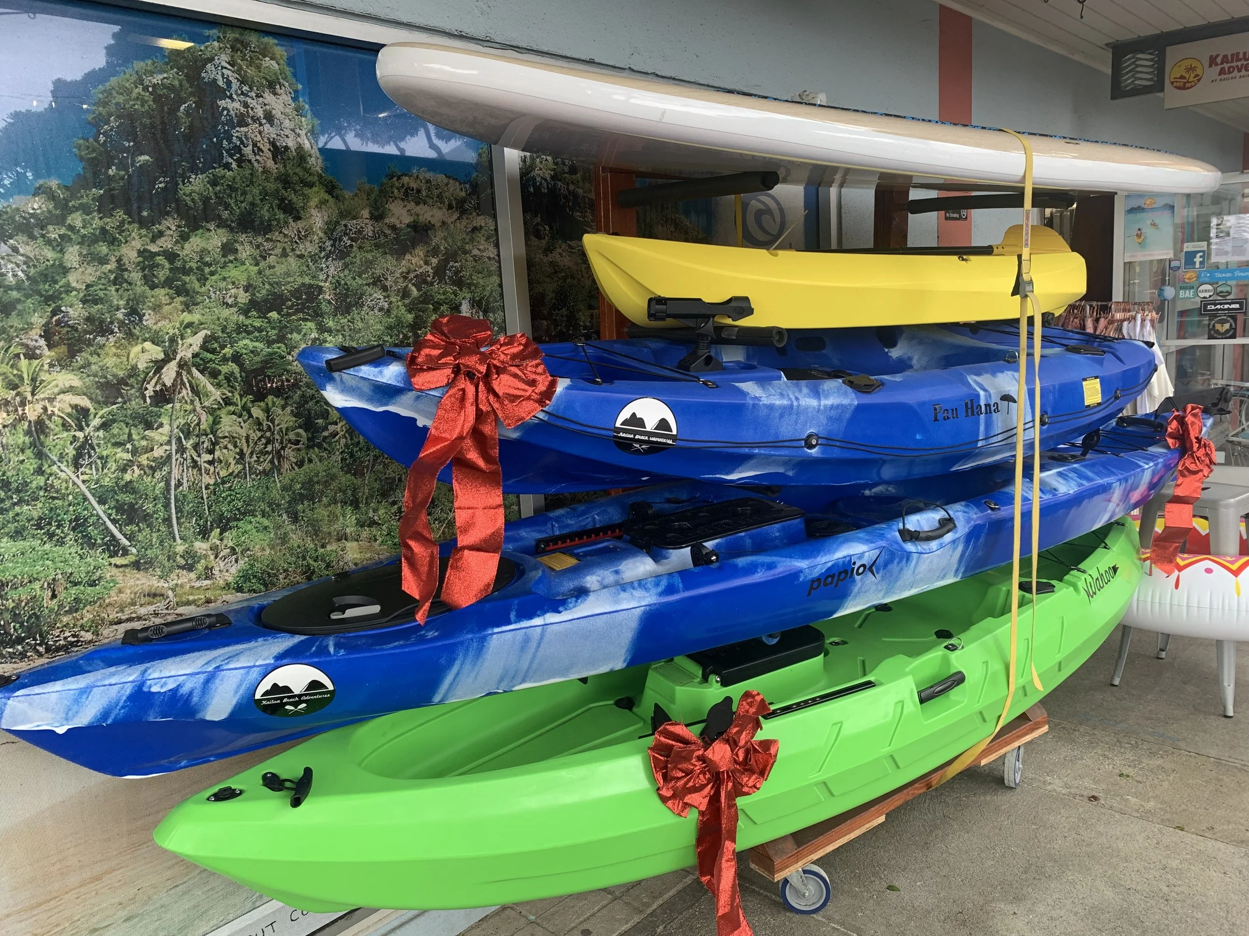 Two kayaks, one yellow and one blue, with red bows attached, stacked on top of two other kayaks, one green and one blue, against a backdrop of a tropical landscape