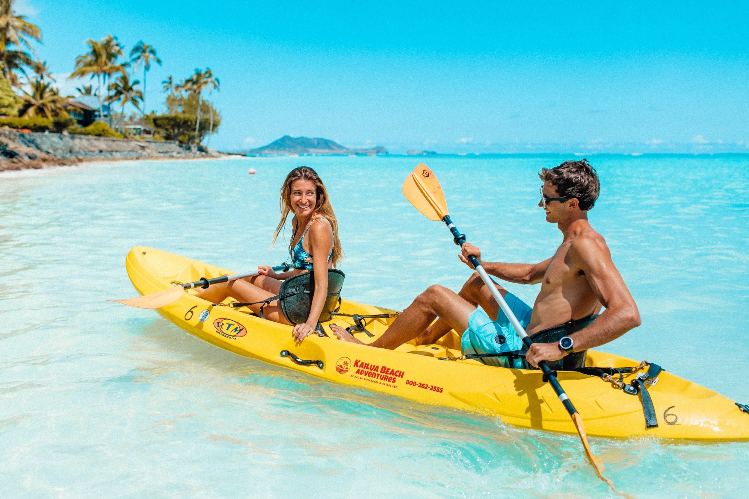 A young man who's holding a paddle and woman looking back at him smiling, kayaking on clear, turquoise water near a beach with palm trees, blue sky, and distant islands.