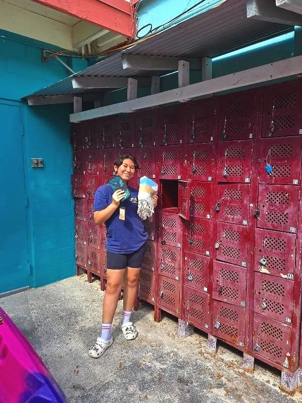 A woman smiling and giving a thumbs up in front of maroon lockers at Kailua Beach Adventures, holding a wallet and towel, wearing a blue t-shirt, shorts, socks, and crocs.