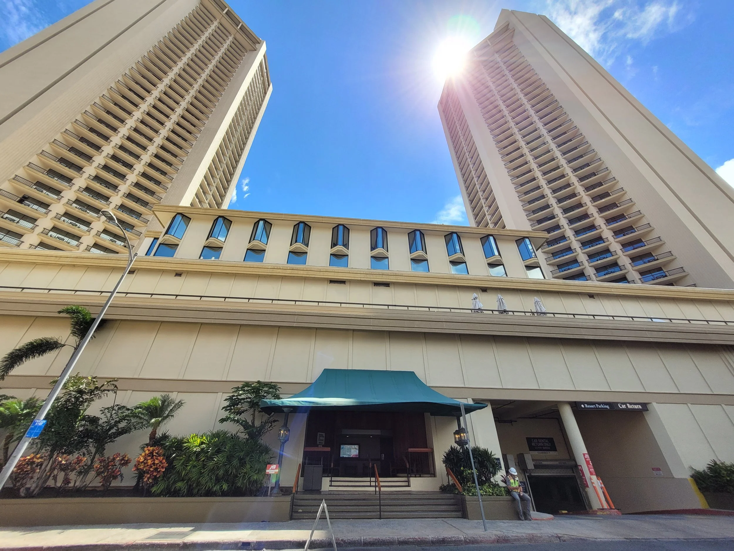Low-angle view of two tall residential or hotel buildings under a bright blue sky with the sun shining. The buildings have numerous balconies and windows, and there are plants and a person sitting near an entrance at the bottom.