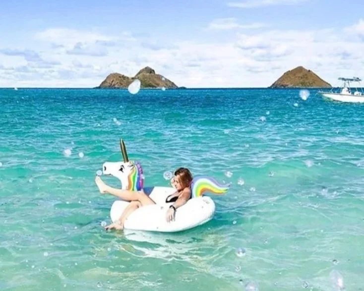 A person floating on a unicorn-shaped inflatable in clear ocean water with small islands in the background and a boat in the distance.