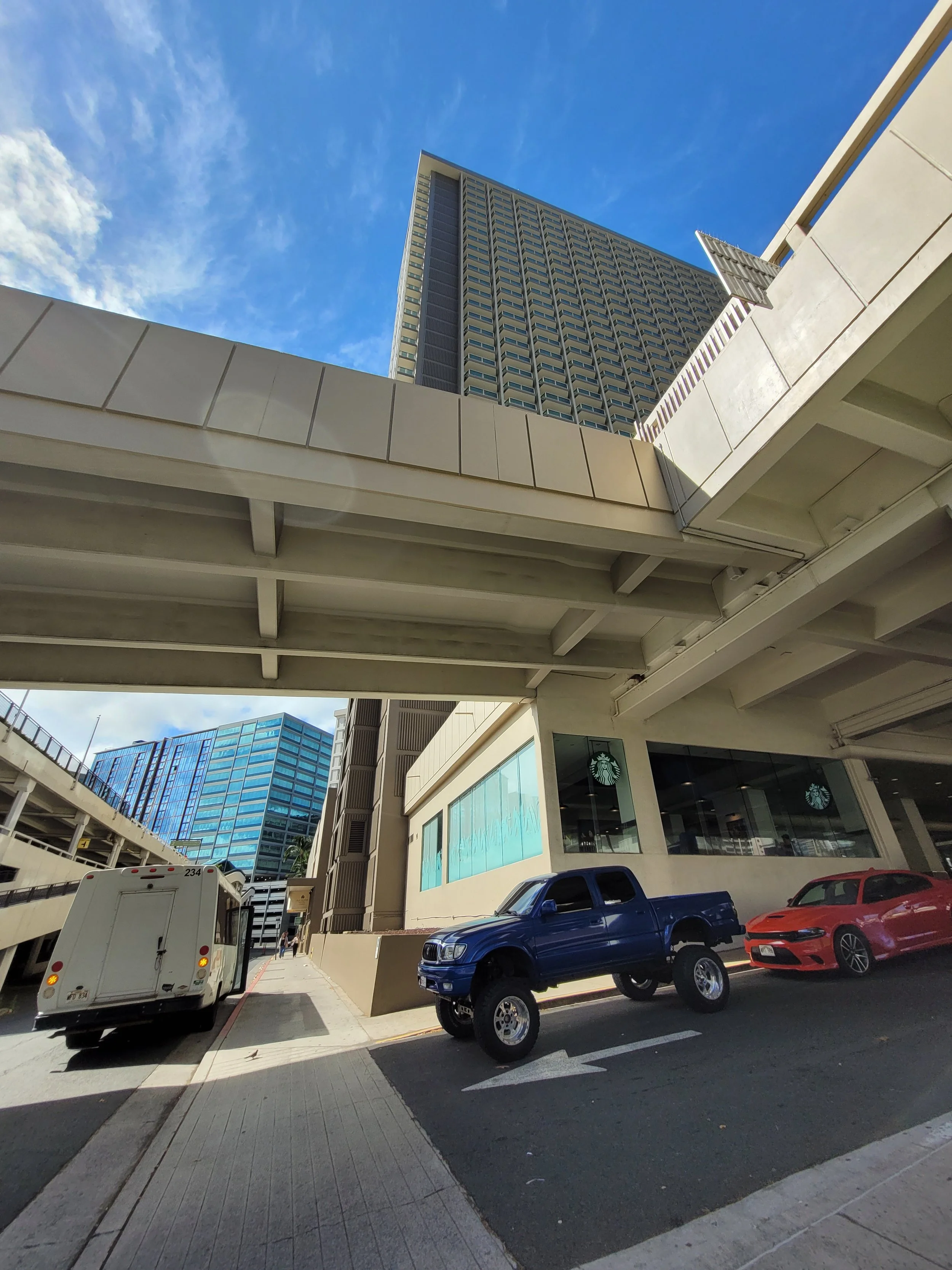View of a city street with a raised highway, modern high-rise buildings, and parked cars, including a blue lifted truck, a red sports car, and a white delivery truck, under a blue sky with clouds.