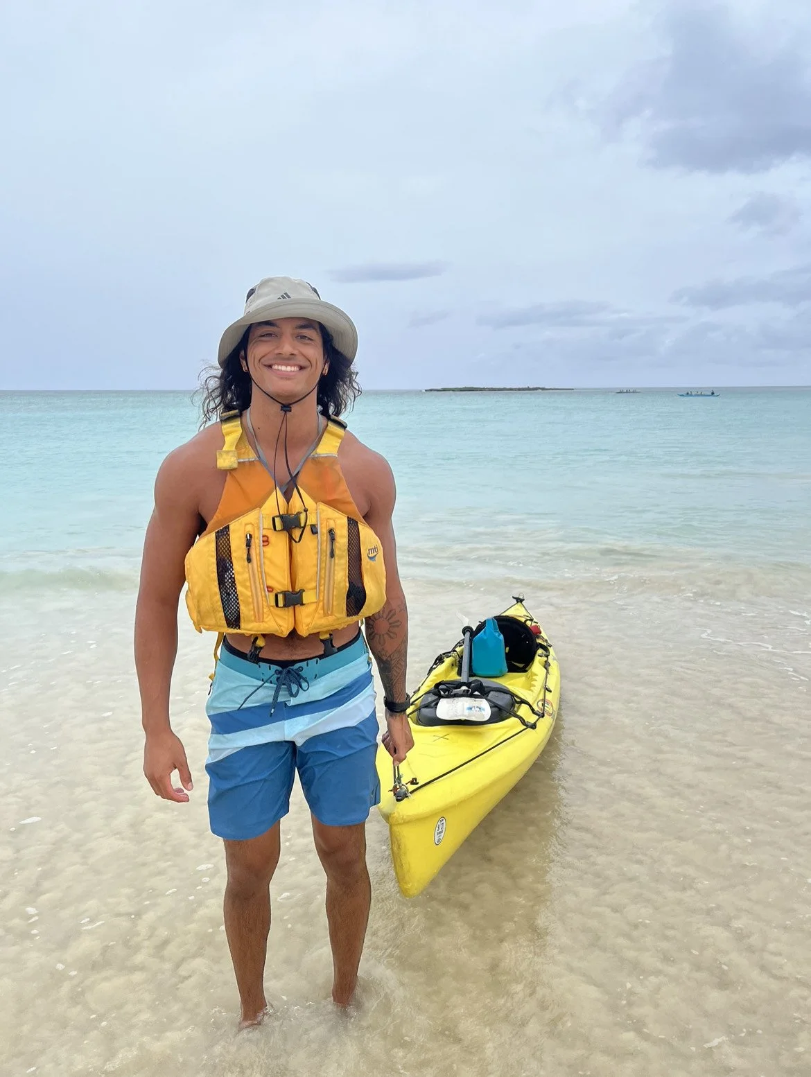 A smiling man standing on a beach next to a yellow kayak, wearing a yellow life jacket, blue shorts, a beige hat, and holding a paddle.