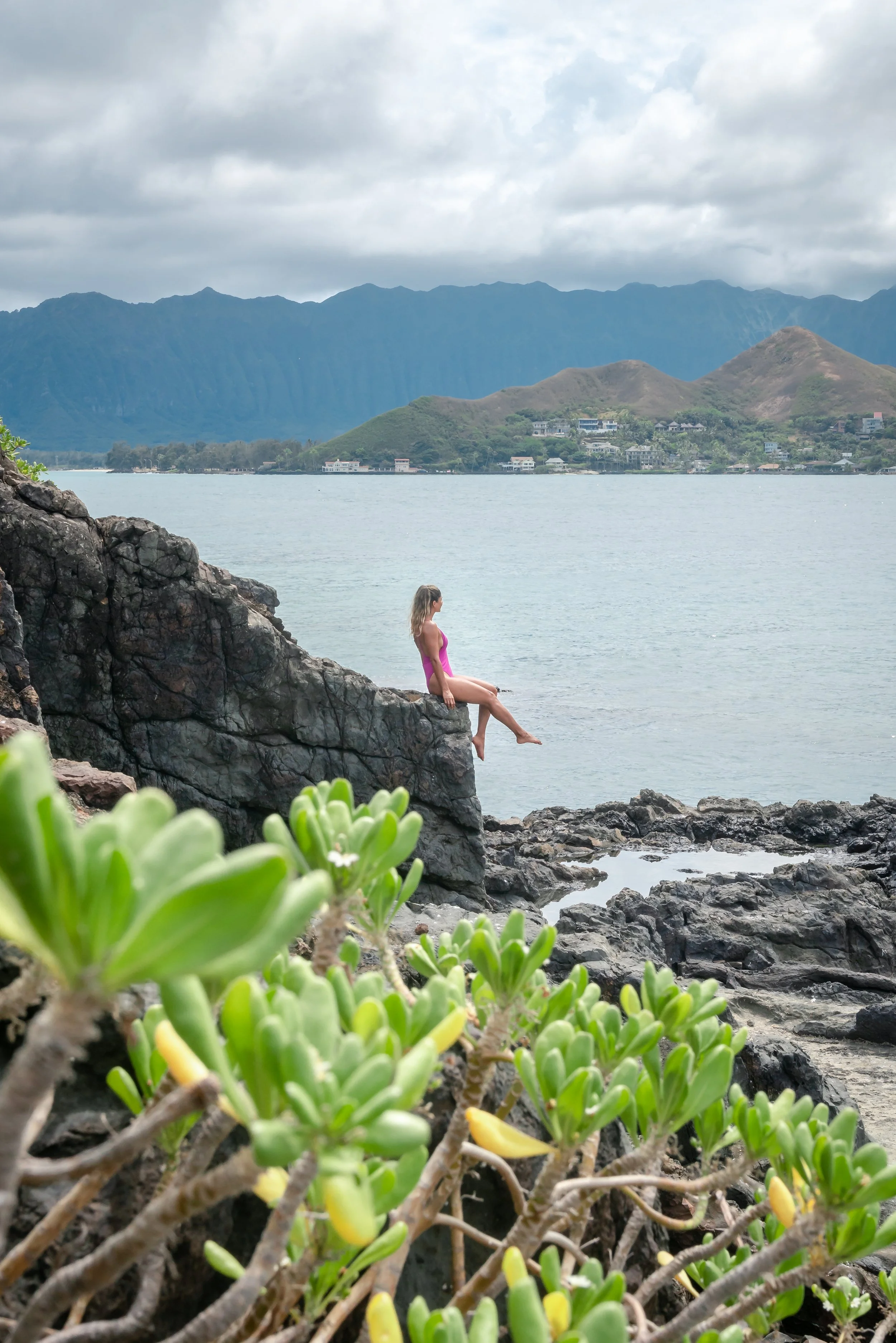 A woman in a pink swimsuit sitting on a rock ledge by the ocean with lush green plants in the foreground and mountains in the background.
