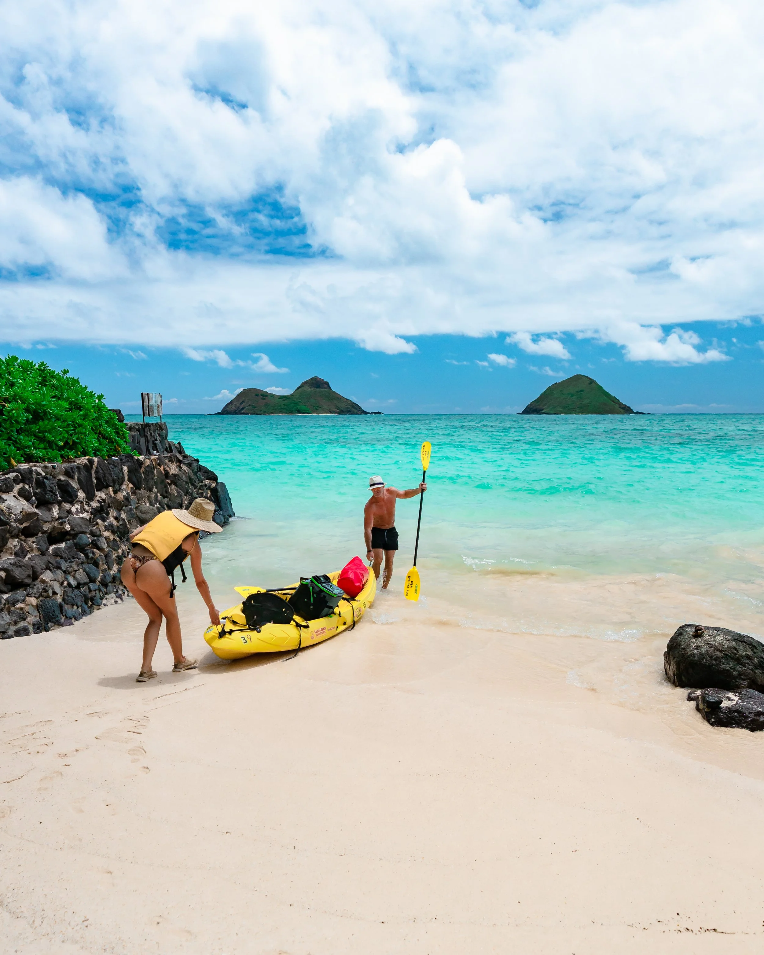 Two people preparing a yellow kayak on a sandy beach, with a tropical ocean, small islands, and partly cloudy sky in the background.