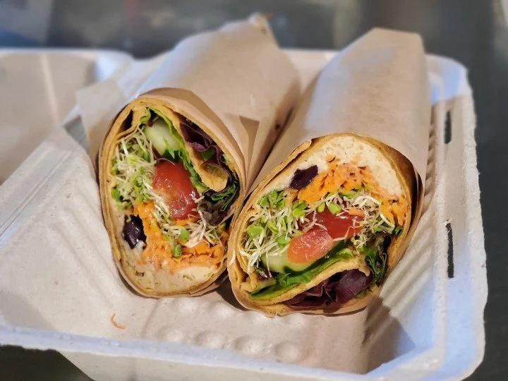 Two veggie wraps cut in half, revealing lettuce, tomato, shredded carrots, sprouts, cucumber, and other fresh vegetables inside a tortilla, served on a paper-lined tray.