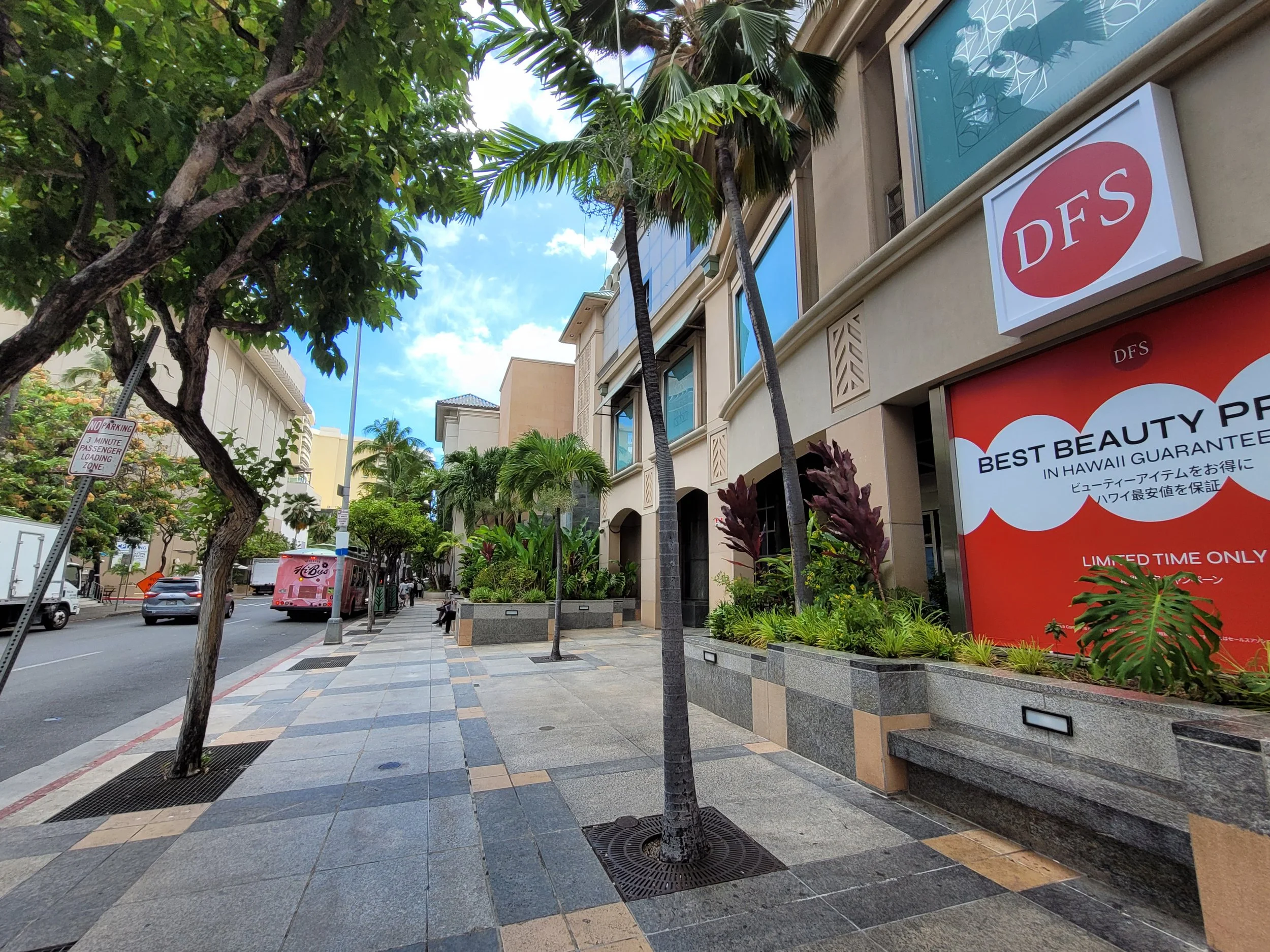 Street scene with trees, a sidewalk with planters, shops with decorative facades, and a moving pink bus in Hawaii.