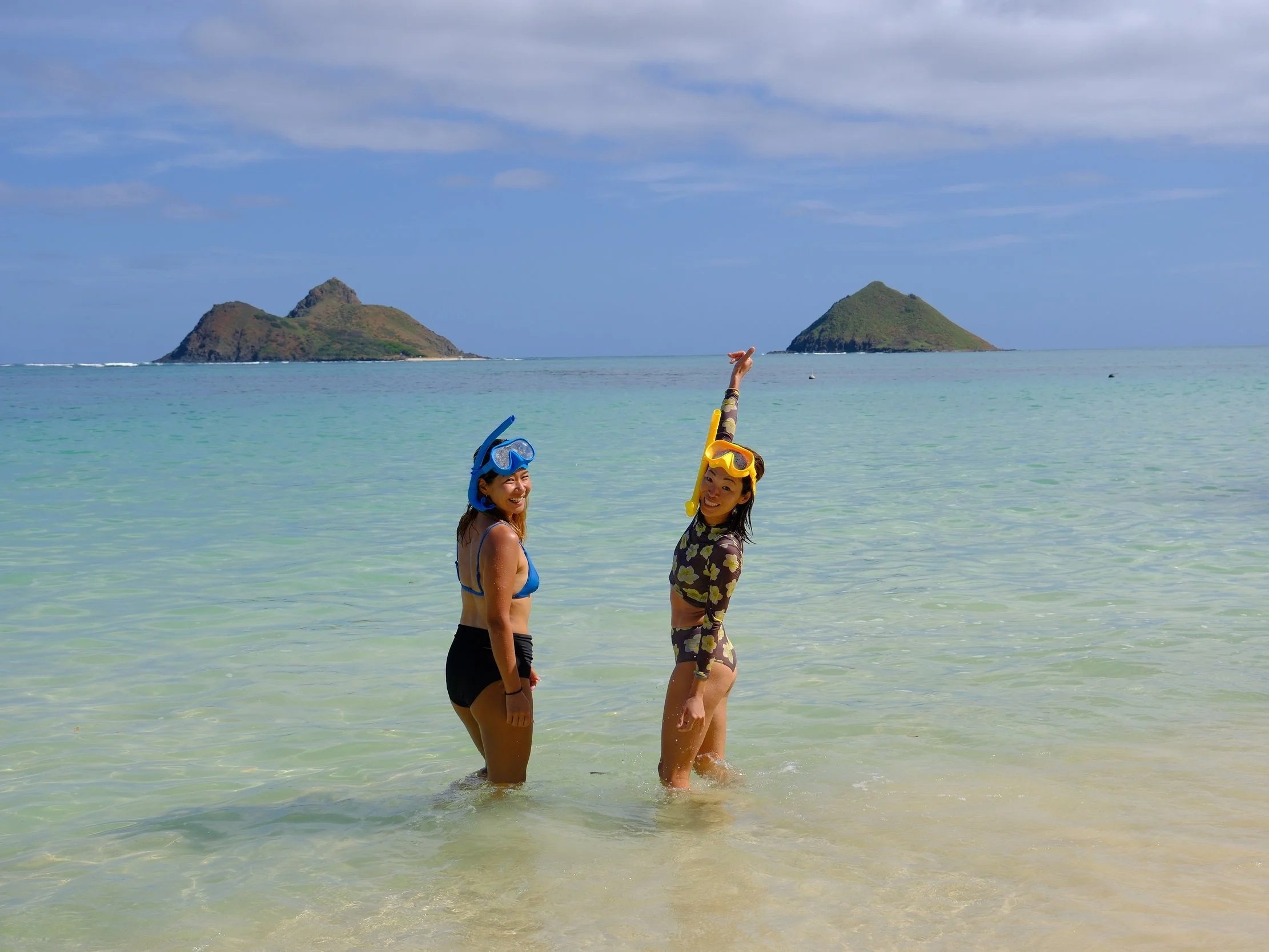 Two women in swimsuits and snorkeling gear lying on a sandy beach with the ocean in the background, smiling at the camera.
