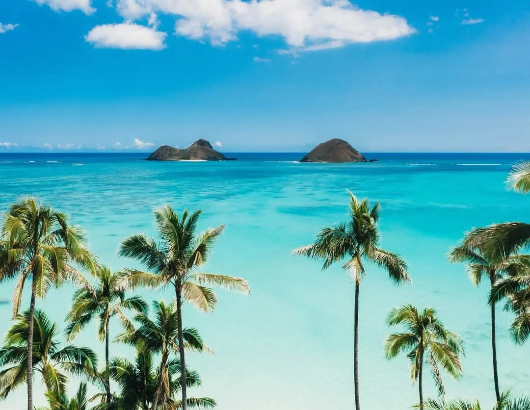 Tropical beach with turquoise water, palm trees in the foreground, and the Mokulua islands in the distance under a blue sky with some clouds.
