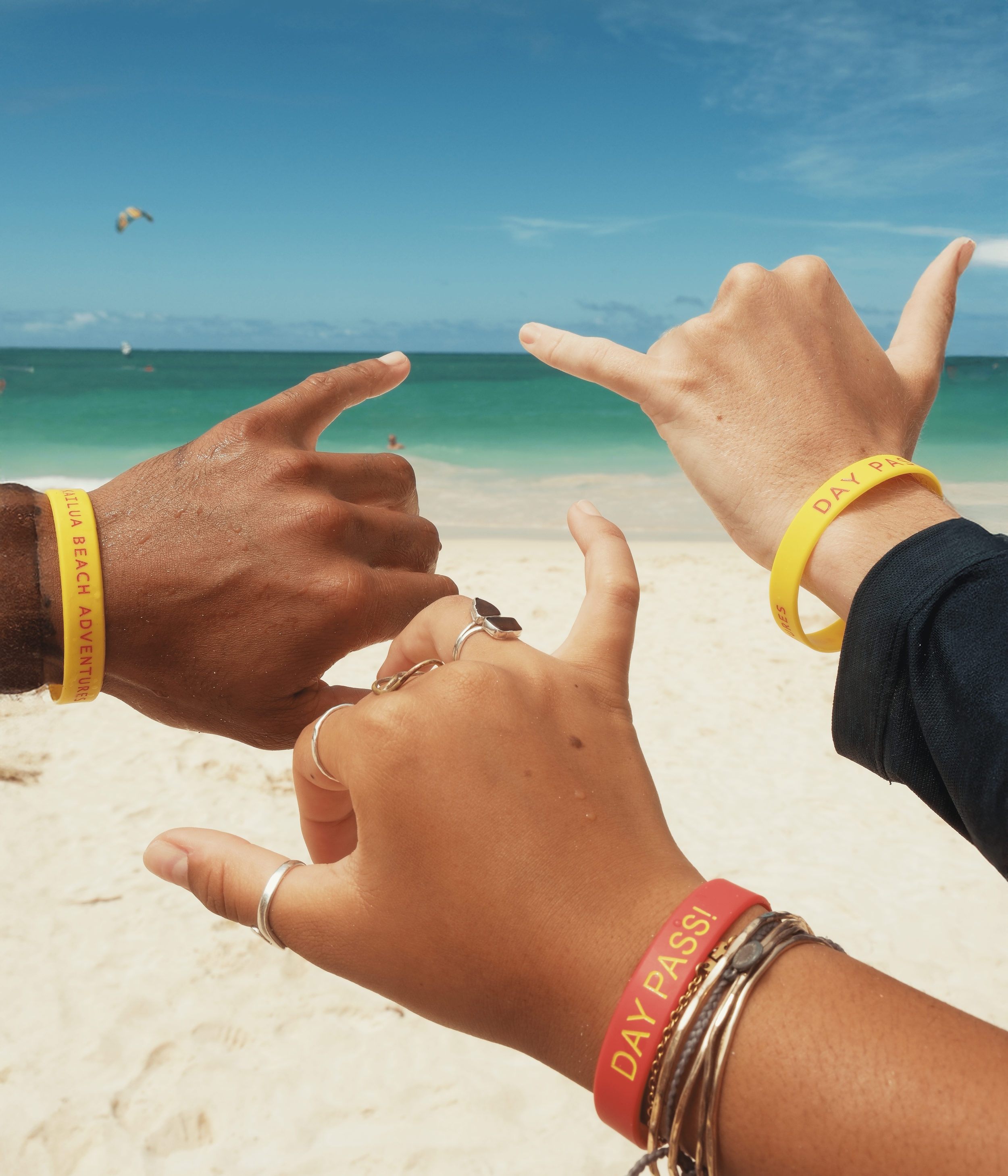 Three hands wearing yellow and red wristbands with the words 'Day Pass!' and 'Kauai Beach Adventure' forming a frame against a beach background with sand, ocean, and blue sky.