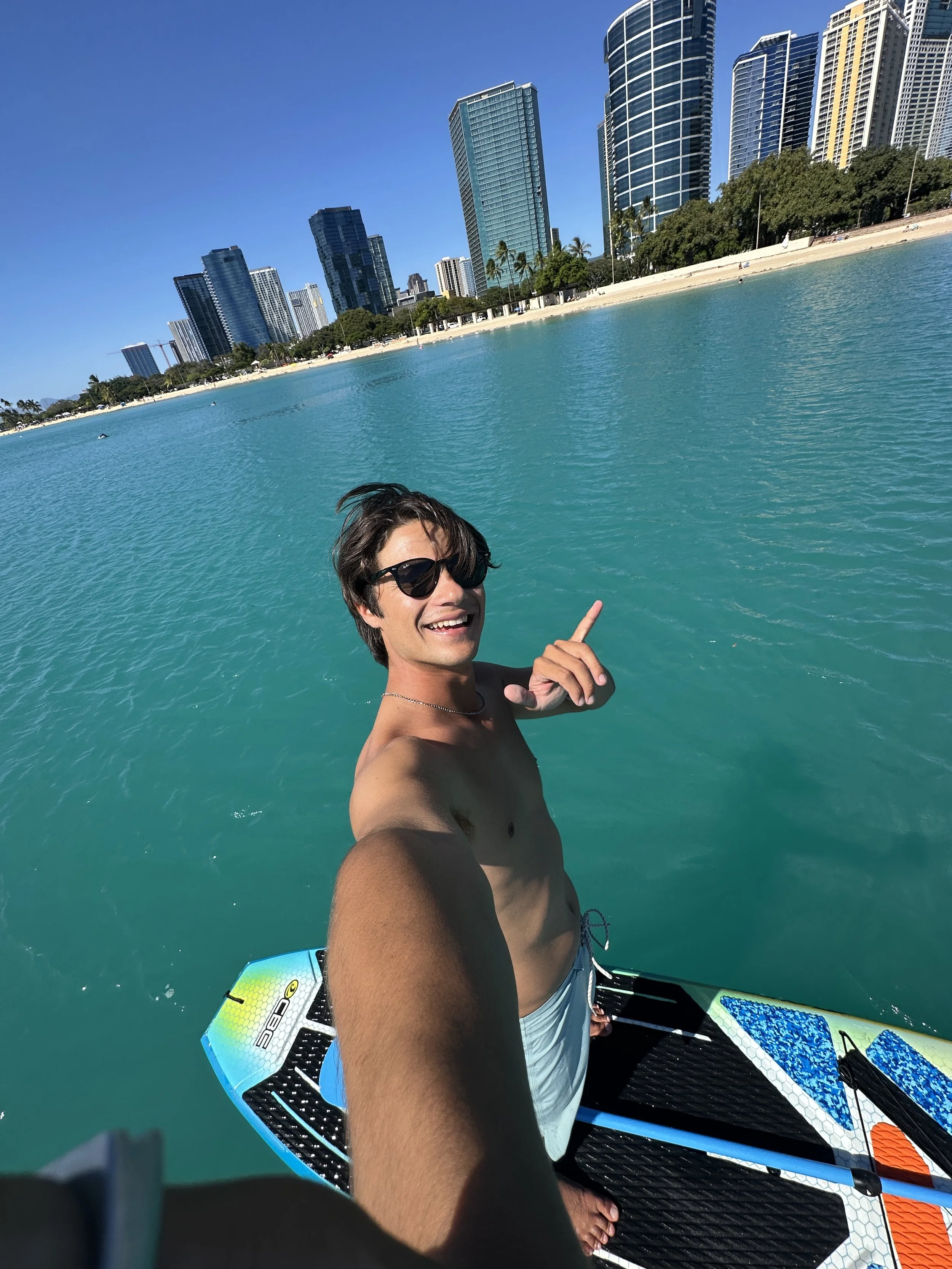 Young man on a paddleboard taking a selfie in turquoise water with a city skyline of tall modern buildings and a sandy beach in the background.