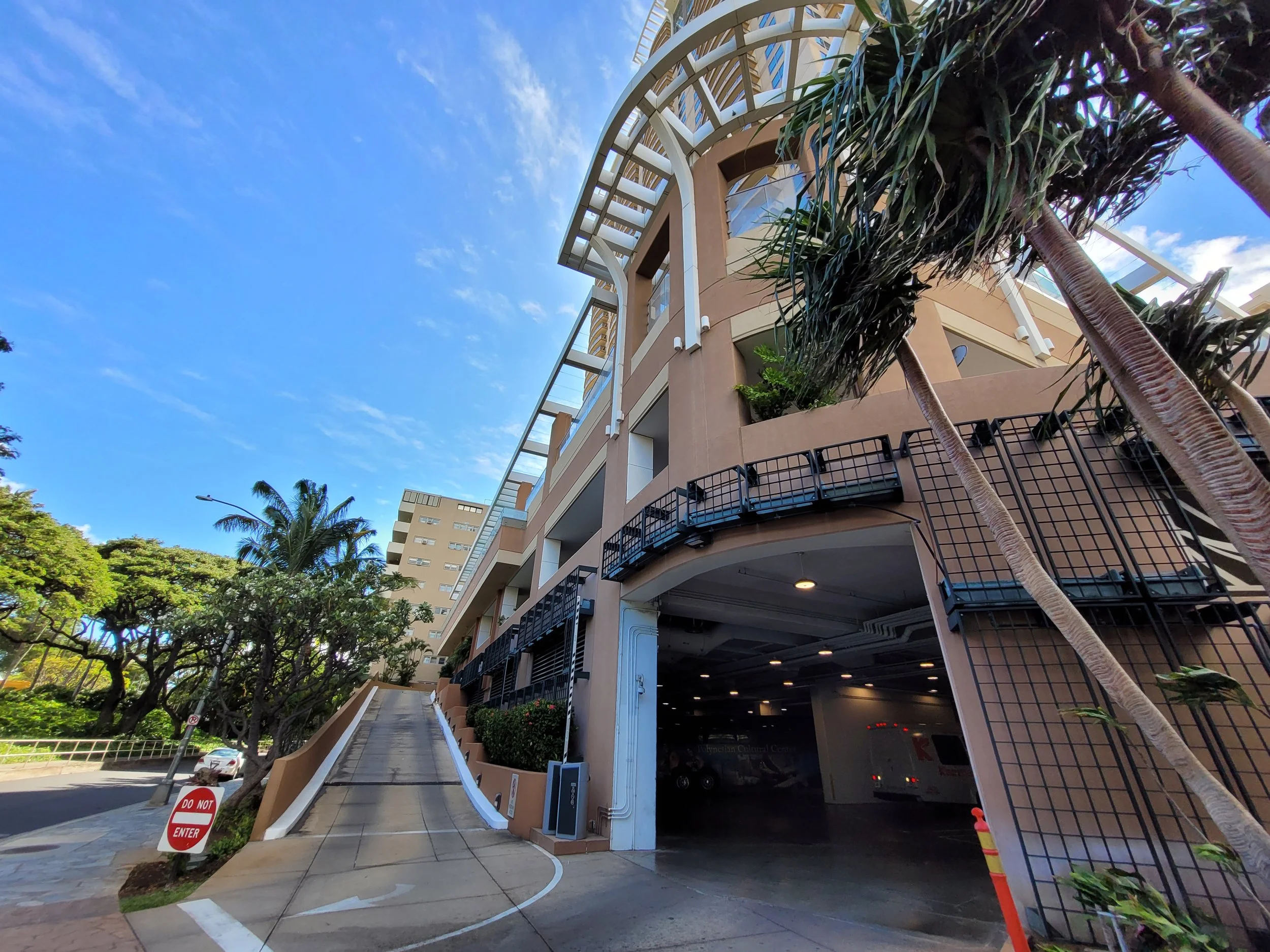 A multi-story building with a parking garage entrance on the ground level, a steep driveway leading up on the left side, and surrounded by tropical trees and palm trees under a blue sky with some clouds.