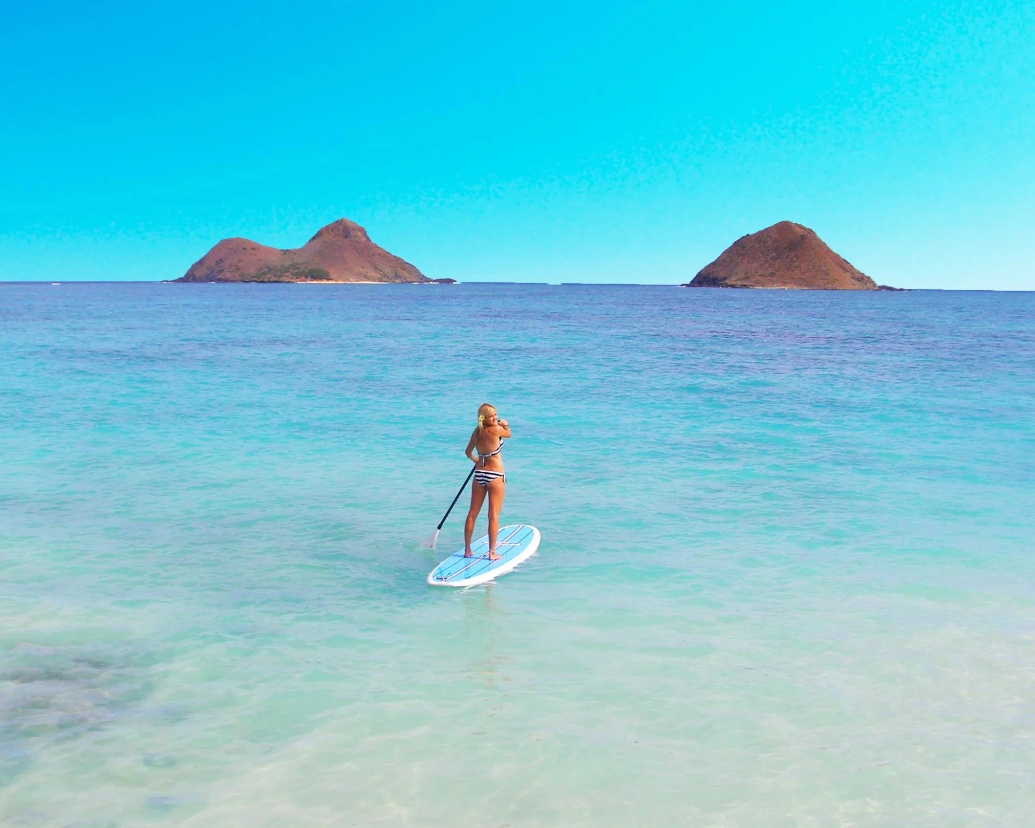 A woman standing on a paddleboard in clear blue waters with two small islands in the background.