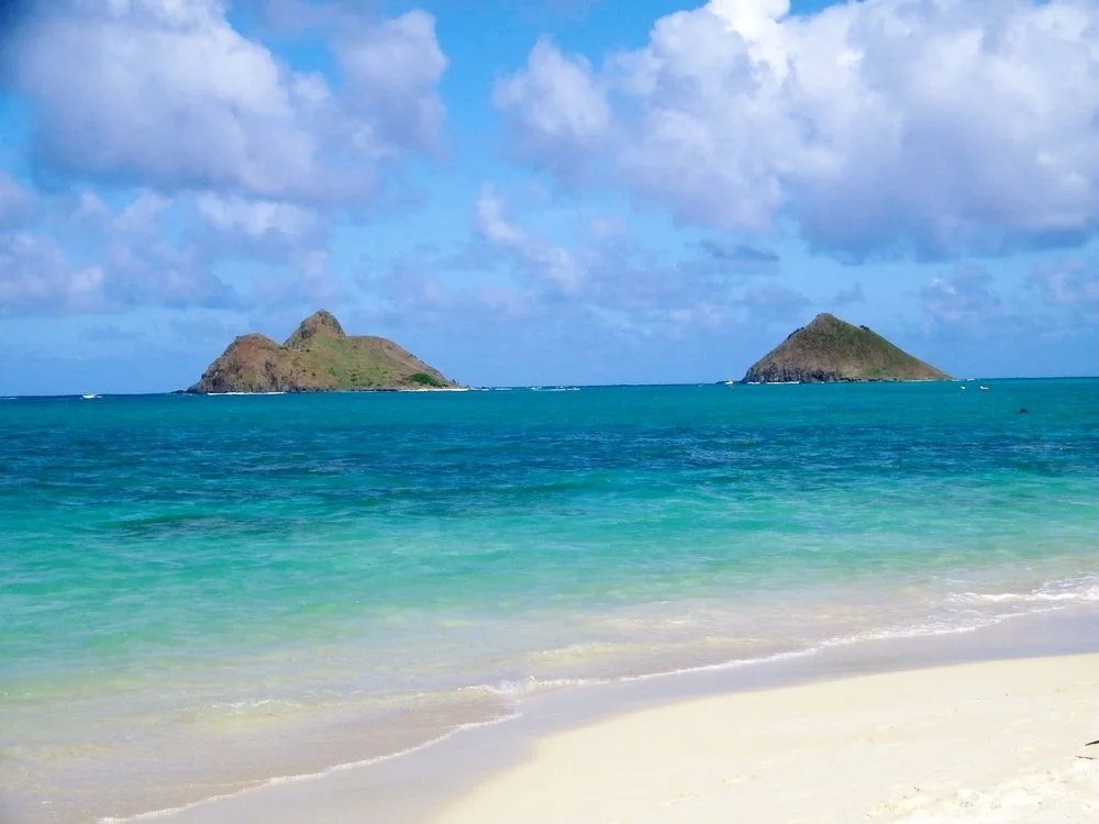 Tropical beach with white sand, turquoise water, and two small islands in the distance under a partly cloudy sky.