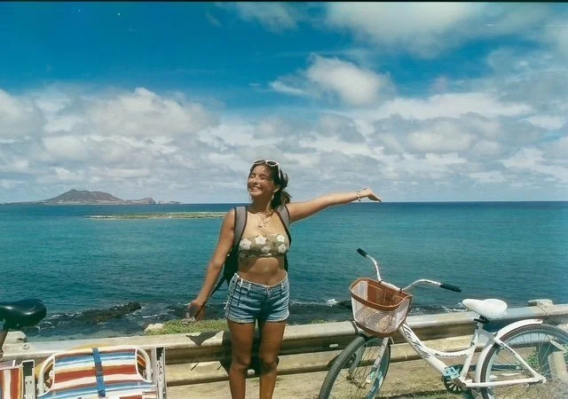 A woman standing by a railing overlooking the beach with her arms outstretched, smiling, wearing sunglasses, a bikini top, and shorts, with bicycles and a beach chair nearby with the ocean in the background.