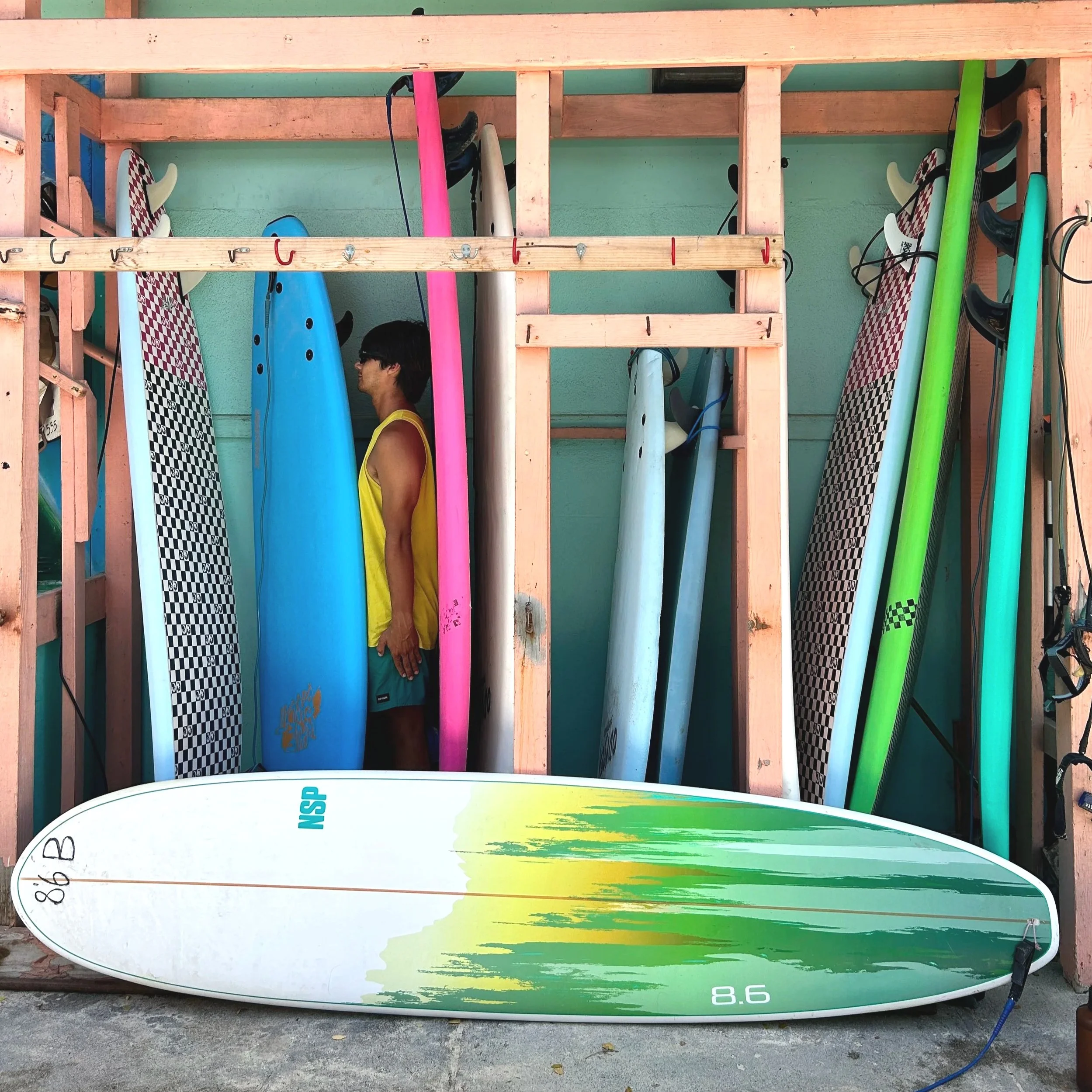 A person in a yellow tank top and blue shorts standing among various colorful surfboards stored vertically in a wooden rack, with a large surfboard lying at their feet.