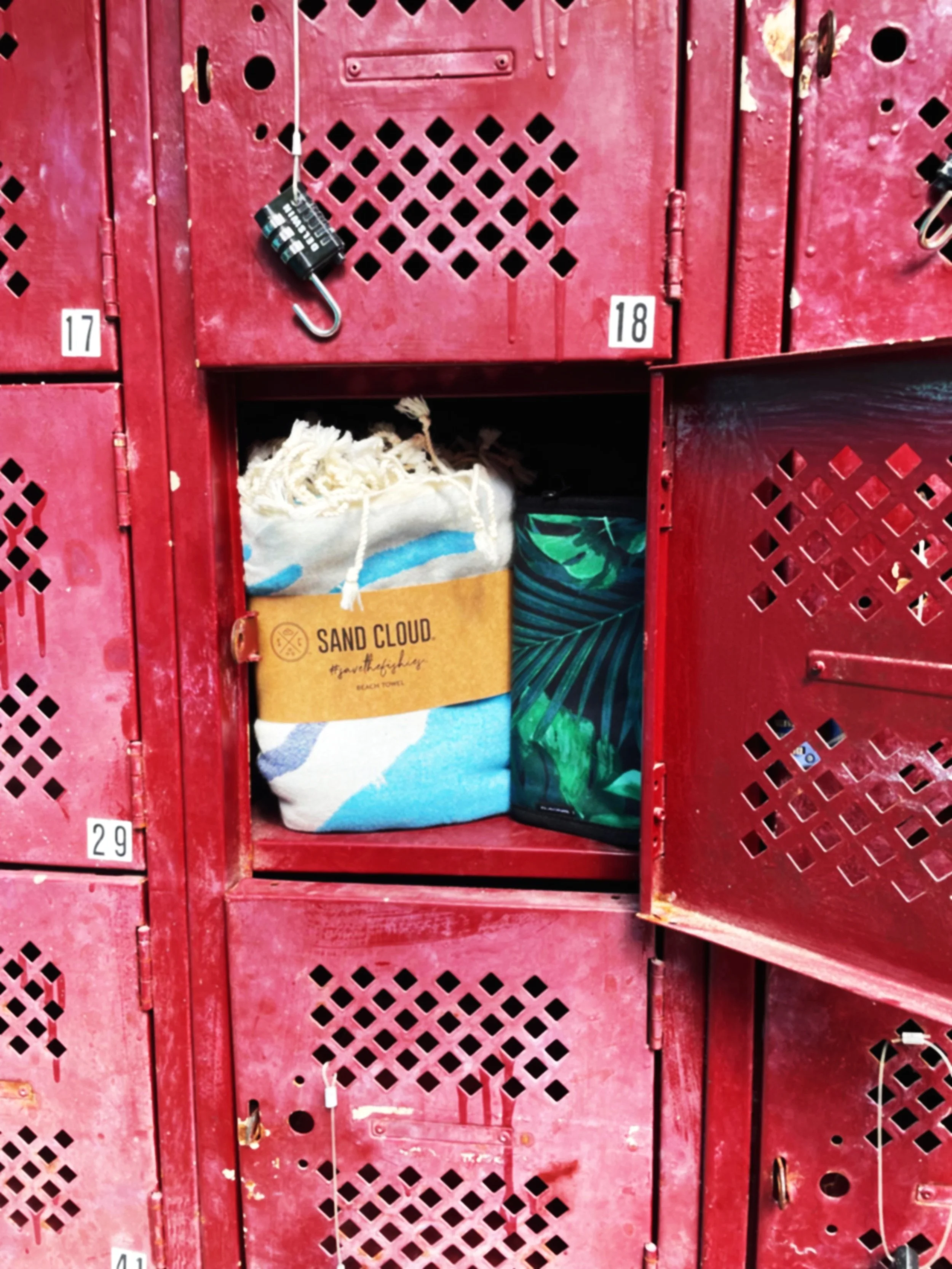 Open red locker containing a folded beach towel labeled 'Sand Cloud' and a green patterned bag.