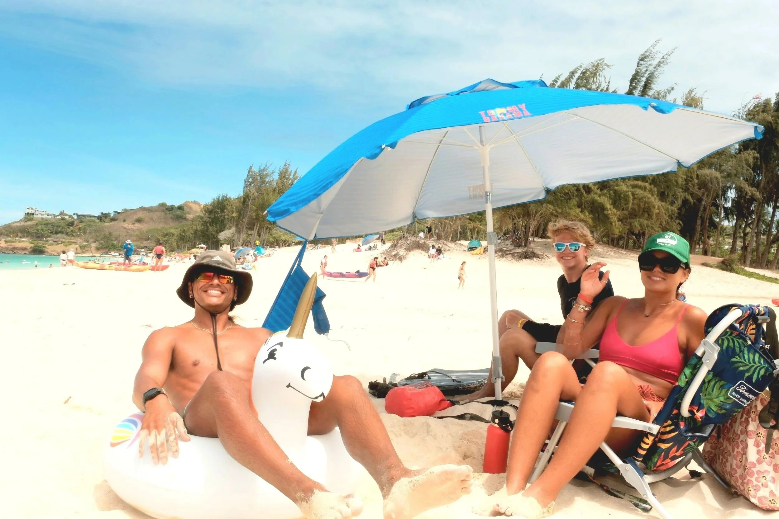 Three friends relaxing on a beach under a blue and white umbrella, with one person sitting on a unicorn-shaped float, all smiling and wearing sunglasses.