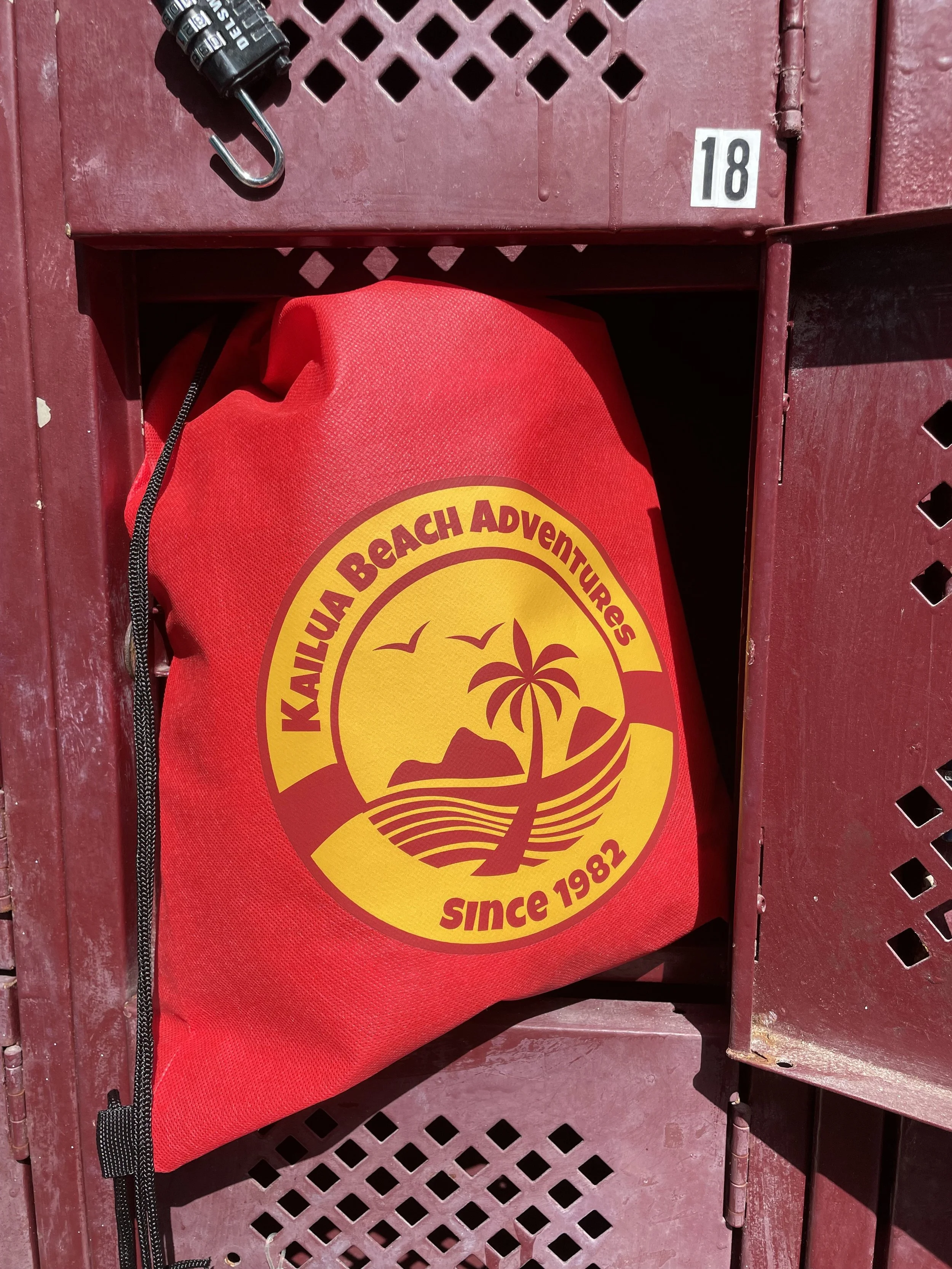 A red dry bag with a yellow and maroon logo reading "Kailua Beach Adventures Since 1982", placed inside a maroon-colored locker.