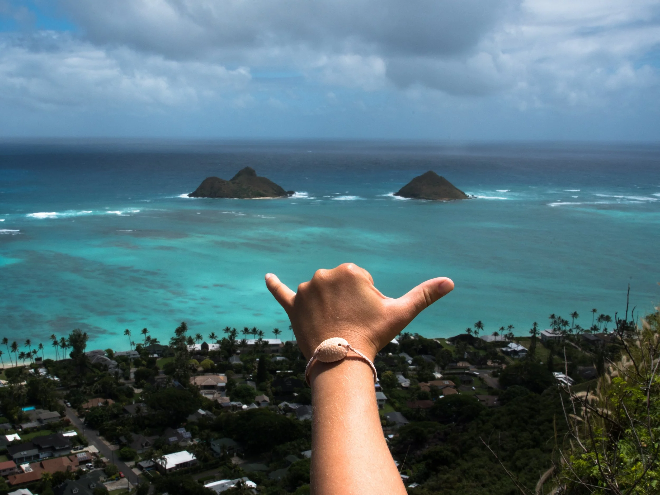 A person is making a shaka sign with their hand, wearing a bracelet with a shell, overlooking a coastal view with turquoise water, Lanikai Beach and the mokulua islands