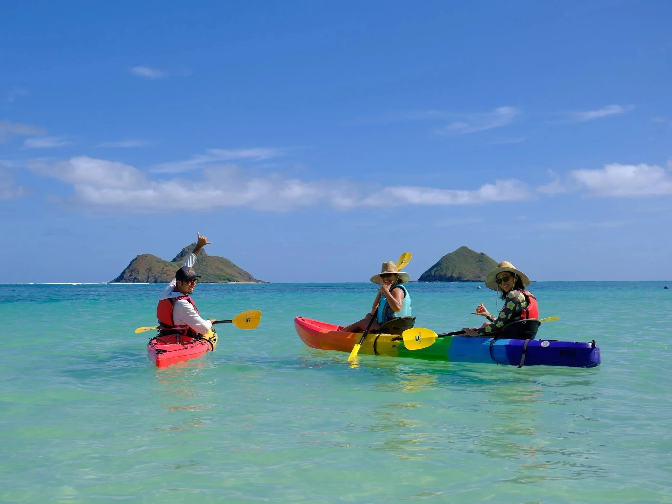 A smiling couple kayaking in clear turquoise waters near a tropical shoreline with palm trees and rocky coast.