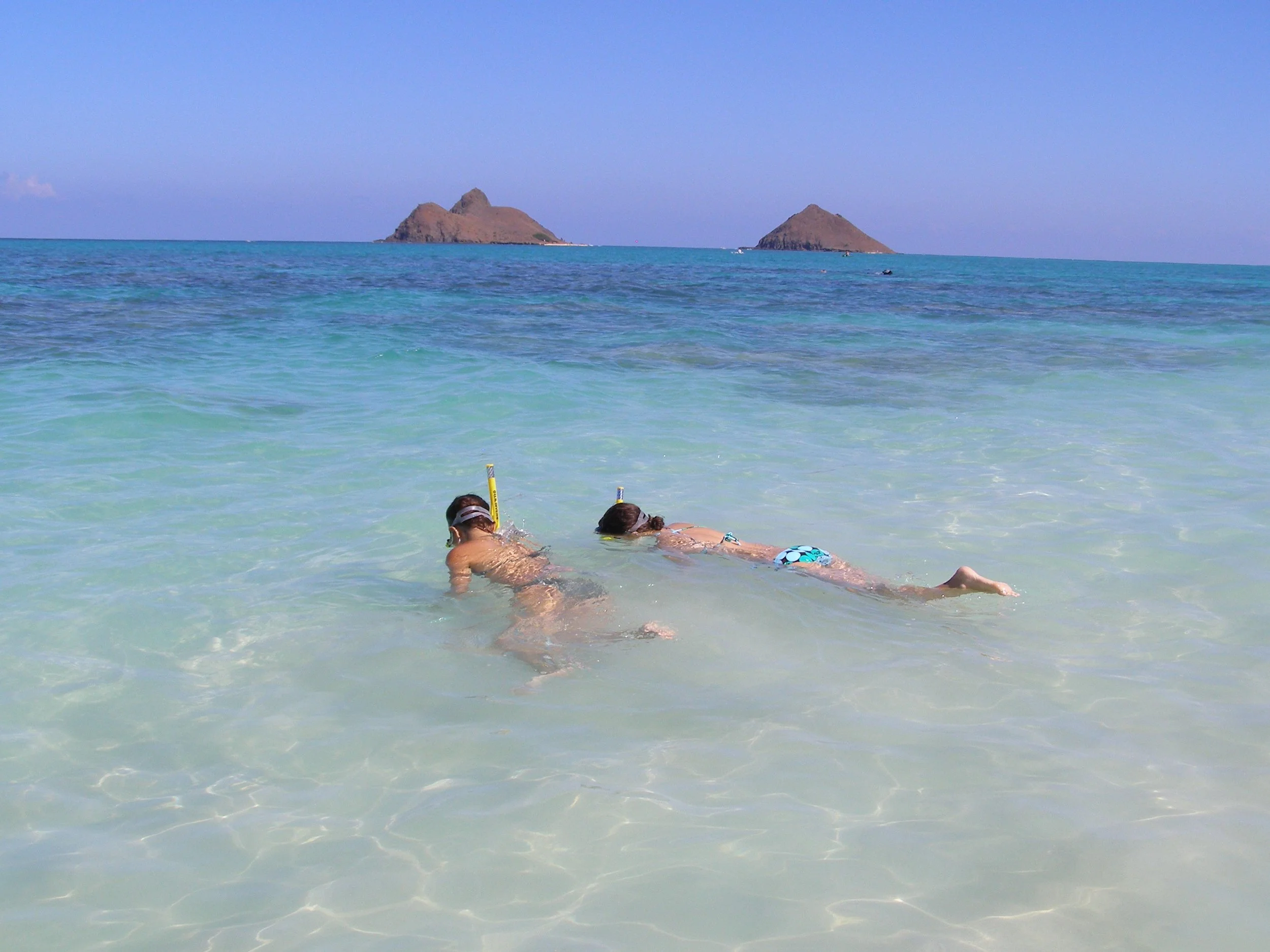 Two children in swimsuits with snorkeling gear explore shallow waters near the beach with greenish water and distant islands.