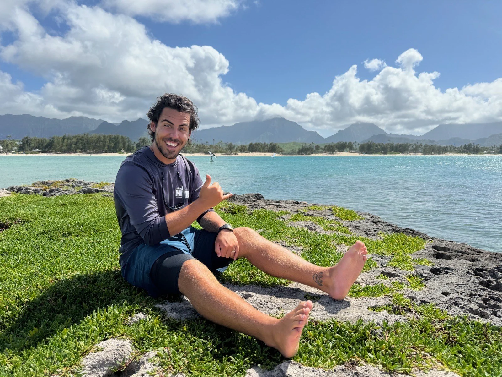Young man sitting on grass by the beach on Flat Island (Popoia), smiling, with mountains and cloudy sky in the background, making a shaka hand gesture.